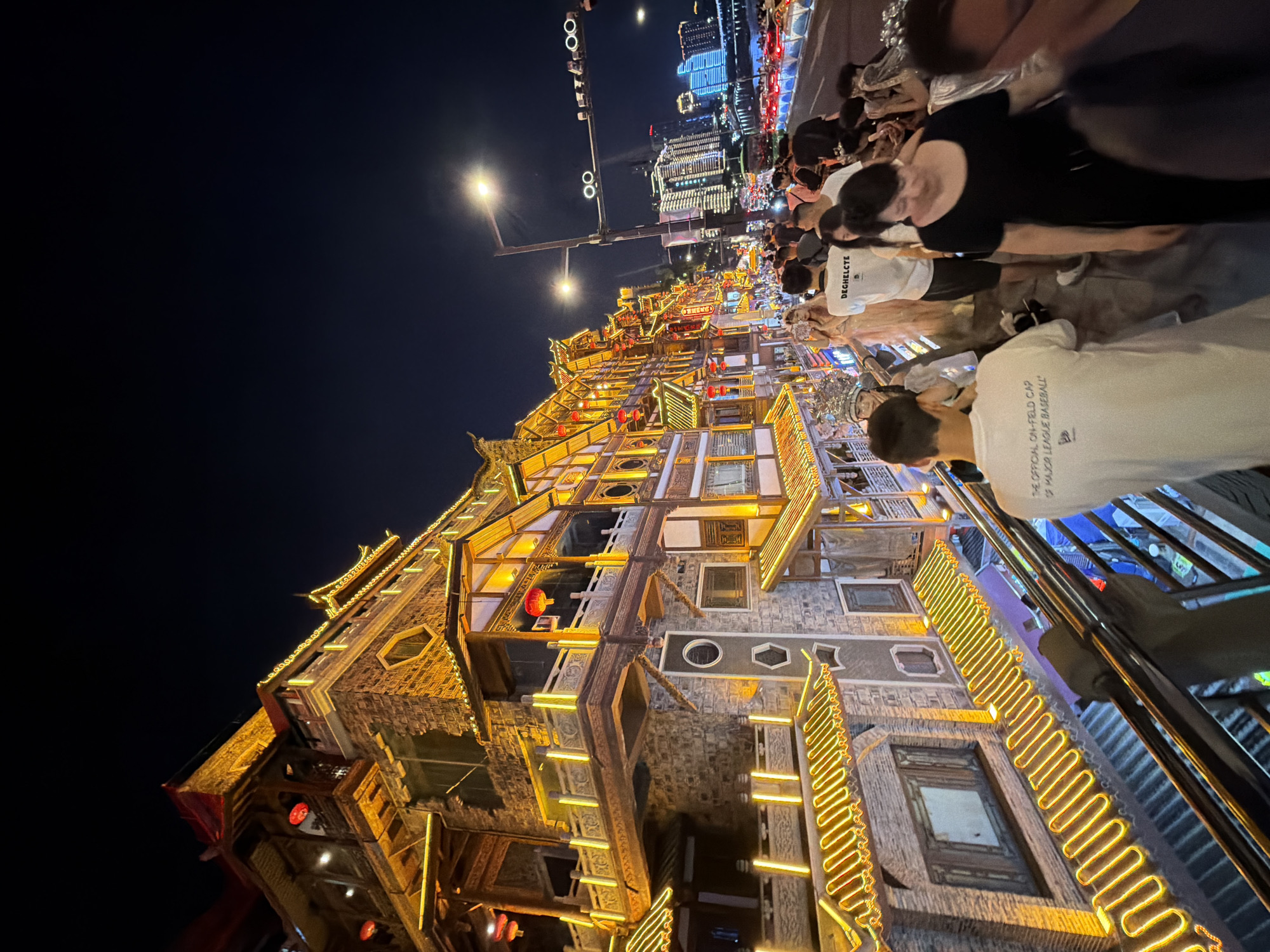 The image depicts a lively nighttime scene on a pedestrian walkway in the Yuzhong District of Chongqing, China, likely near the Hongyadong complex. The architecture is characterized by traditional Chinese-style multi-story buildings, illuminated with vibrant yellow neon lights that accentuate the eaves and decorative elements. Red lanterns are visible, adding to the festive atmosphere. A large crowd of people are walking along the elevated walkway, suggesting a popular tourist or entertainment area. Some individuals are dressed casually, while others appear to be in more formal attire. In the background, the cityscape is visible with modern high-rise buildings and illuminated bridges over a body of water, indicating Chongqing's urban environment. The overall impression is one of a bustling commercial and cultural hub at night.