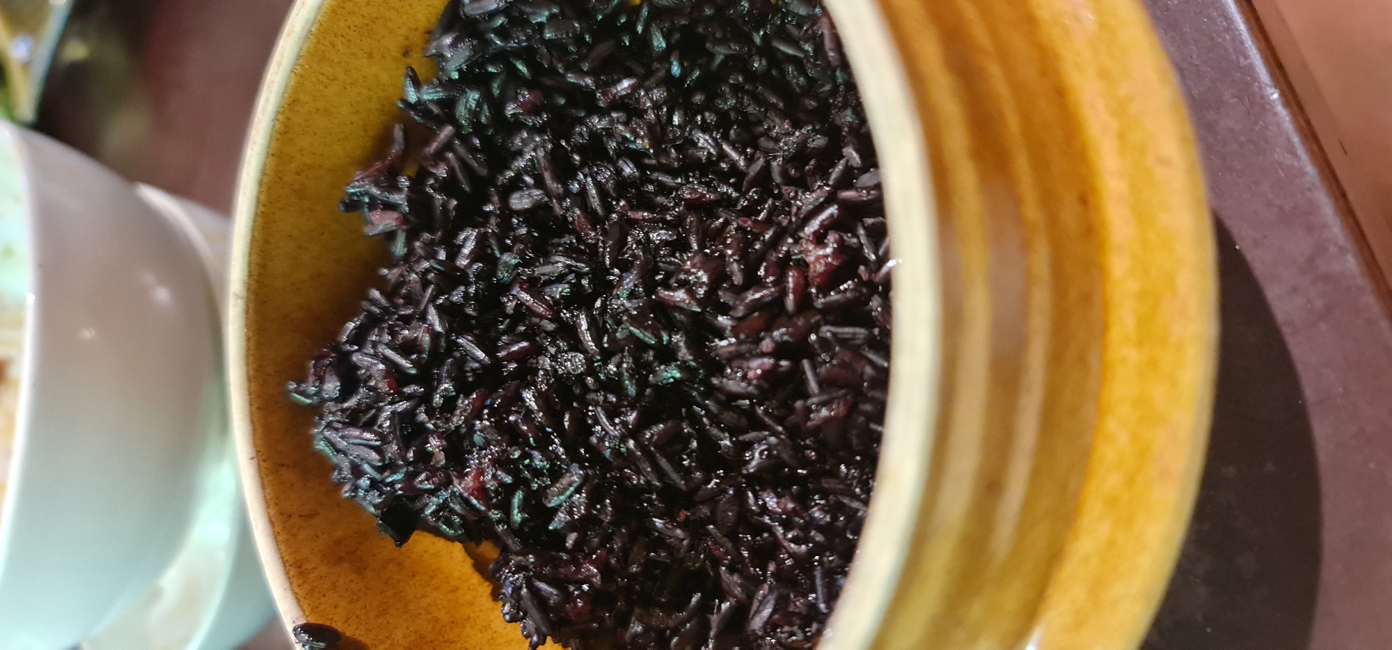 A bowl of black rice is prominently featured, filling most of the frame. The bowl is yellow with a ribbed texture. The black rice appears glistening and slightly moist. In the background, partially visible on the left, is a white dish. The scene is set on a dark-colored surface. It is unspecified if this image was captured in the city of Đà Nẵng, Vietnam.
