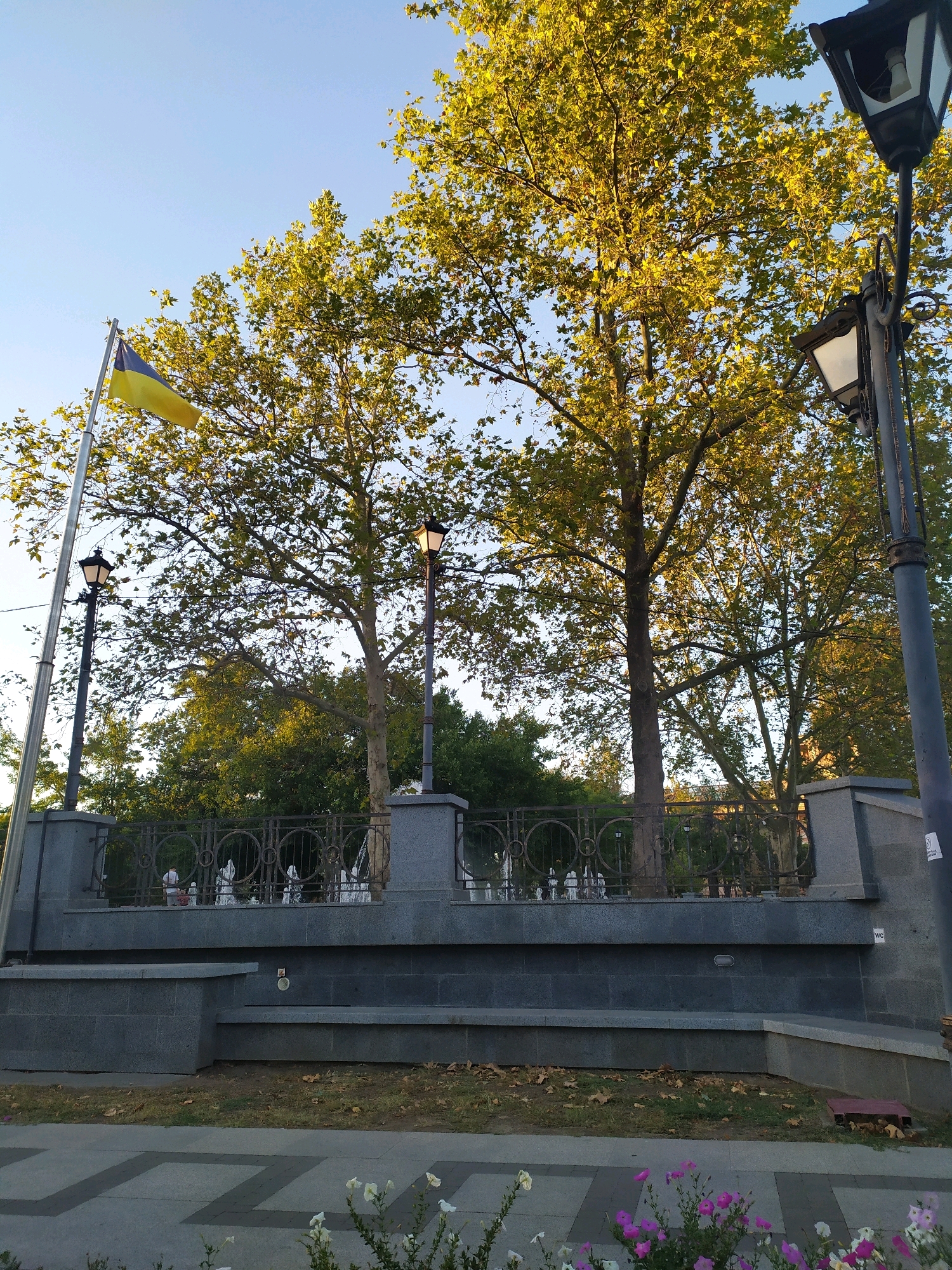 In Mykolaiv, Ukraine, the image captures a sunlit scene featuring a Ukrainian flag waving on a flagpole under a clear blue sky. Tall trees with lush, yellow-green foliage dominate the background, suggesting an autumn setting. In the foreground, a grey stone structure with a decorative metal fence sits behind a paved walkway. Two ornate lampposts stand on either side of the central part of the structure, one on the left and another on the right. The lower part of the image shows a sidewalk with a geometric pattern and small flowering plants, some with white and pink petals. The overall impression is of a well-maintained public space, possibly a park or a memorial site, bathed in natural light.