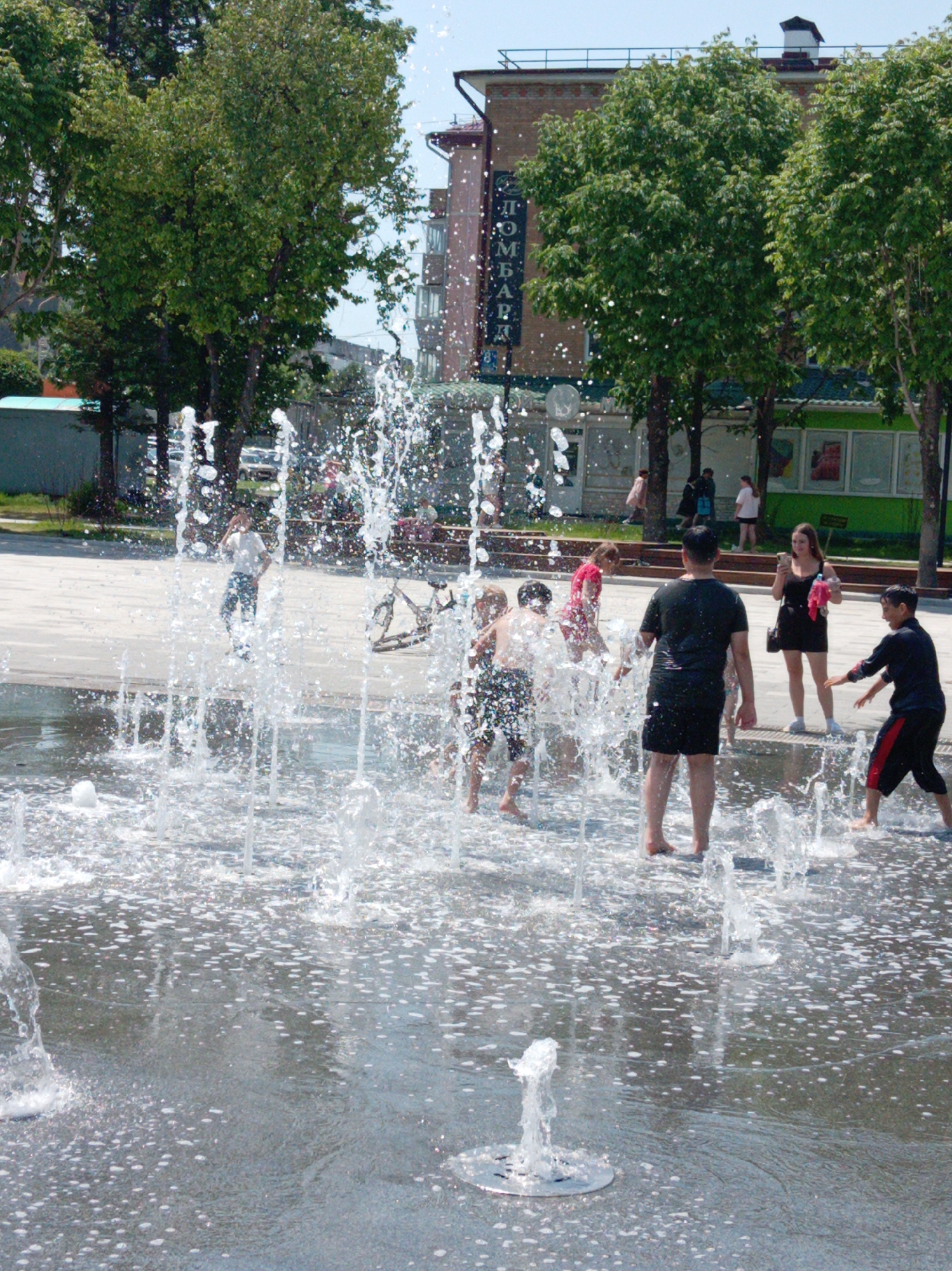 This image captures a vibrant outdoor scene in Artyom, Russia, depicting people enjoying a public ground-level fountain on a bright, sunny day. The foreground and midground are filled with vertical jets of water spraying upwards from the fountain, creating a dynamic splash pad. The paved ground surrounding the fountain is wet and glistening with numerous water droplets.
Several individuals are actively engaged with the fountain. Multiple children, some barefoot and others in swimwear or light clothing, are seen playing directly within the water, running and splashing. An adult male, dressed in a black t-shirt and shorts, also stands in the fountain. At the edge of the water, a young woman in a black dress stands holding a smartphone and a pink item of clothing, observing the scene. In the background, other people are visible walking or standing near the trees and buildings.
The setting appears to be an urban park or public square. Lush green trees line the background, with buildings visible behind them. On one of these buildings, a vertical sign displays the Cyrillic text "ДОМБАРЪ" (DOMBAR). The clear sky and bright lighting indicate it is midday or early afternoon, suggesting warm weather. The overall activity conveys a sense of fun, refreshment, and leisurely enjoyment in a public recreational space.