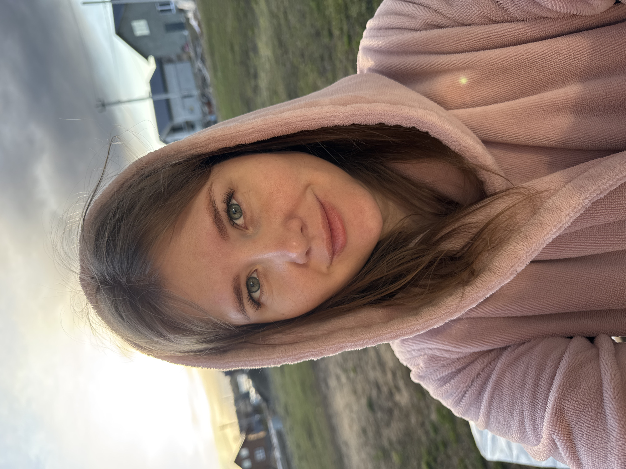 The image is a selfie taken by a fair-skinned woman wearing a pink hooded robe. She has light brown hair and green eyes. The background shows a grassy area and buildings, likely located in the city of Ферма, Russia. The sky is overcast.