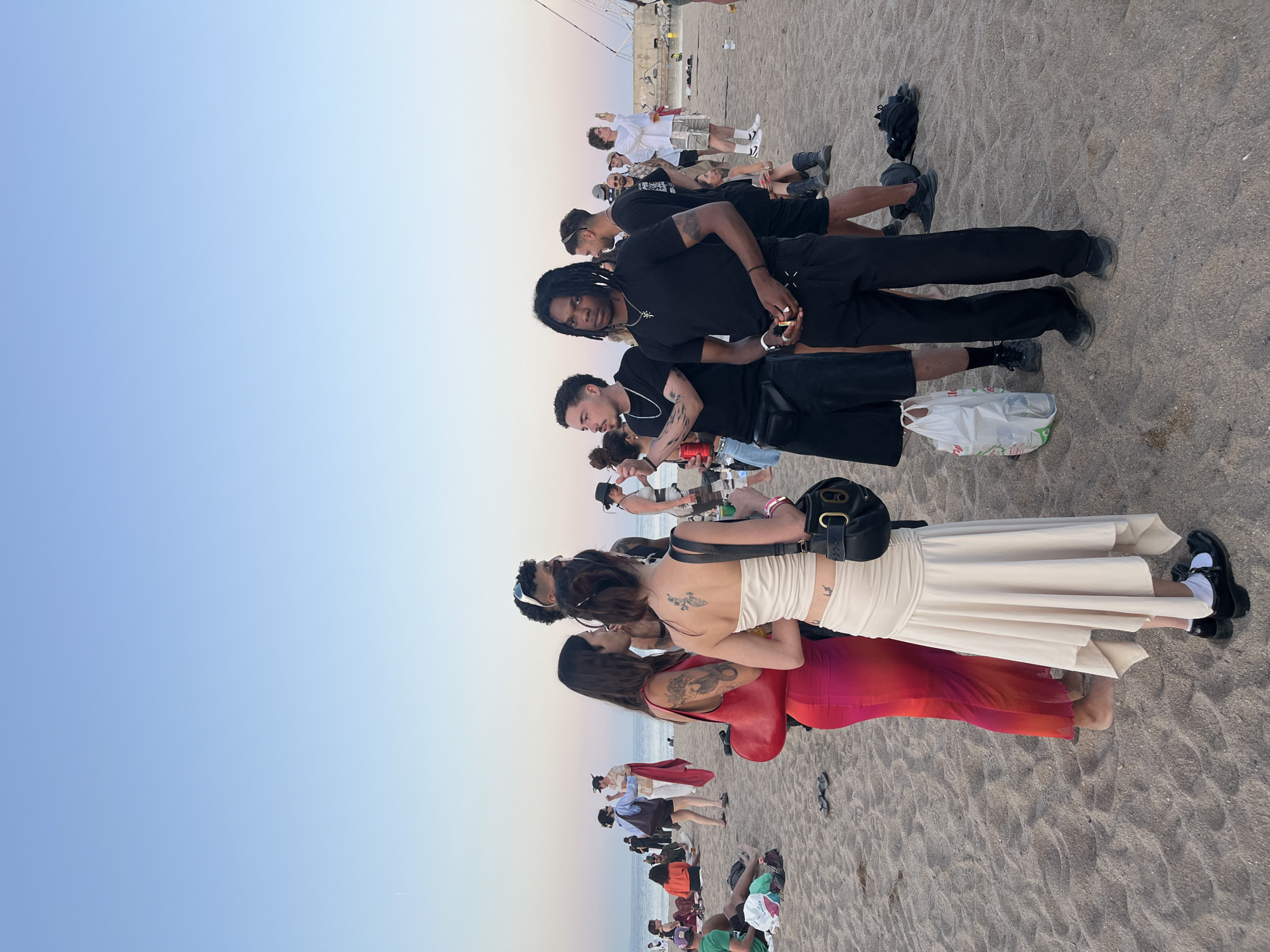 The image captures a beach scene at dusk in Sant Adrià de Besòs, Spain, with a clear sky transitioning from light blue at the top to pale orange and pink near the horizon over the sea. In the immediate foreground on the left, two women are engaged in conversation. The woman on the left wears a sleeveless, form-fitting dress that gradients from red to pink, displays tattoos on her left arm, and carries a red crescent-shaped shoulder bag. Her feet are bare. The woman on the right has her dark hair pulled back, wears a light beige strapless top with a matching ruched, asymmetrical skirt, and exhibits tattoos on her upper back and lower left hip. She carries a black structured shoulder bag and wears black Mary Jane style shoes with white socks. A pair of black flip-flops is visible on the sand behind them.
To the right of the women, a group of three men stands. The leftmost man, with curly dark hair and tattoos on his left arm, wears a black t-shirt, a silver chain, and a black fanny pack, holding a red beverage can. The central man, with dark dreadlocks, wears a black t-shirt, black trousers, a silver chain with a cross pendant, and multiple rings, holding a small object in his hands. The man to his right, with short dark hair and sunglasses perched on his head, wears a black t-shirt and dark shorts, showing tattoos on his right arm. A white plastic shopping bag with green text rests on the sand near these men.
In the midground, several individuals are seated or lounging on the sand on the left, with one person in a green t-shirt visible. On the far right midground, a white mast-like structure is partially visible. Further into the background, numerous people are scattered along the sandy beach, some near the water's edge, including one individual in a white shirt and light shorts holding a phone up, seemingly taking a photograph. The overall environment is a sandy beach with the sea extending to the horizon under a gradient sky.