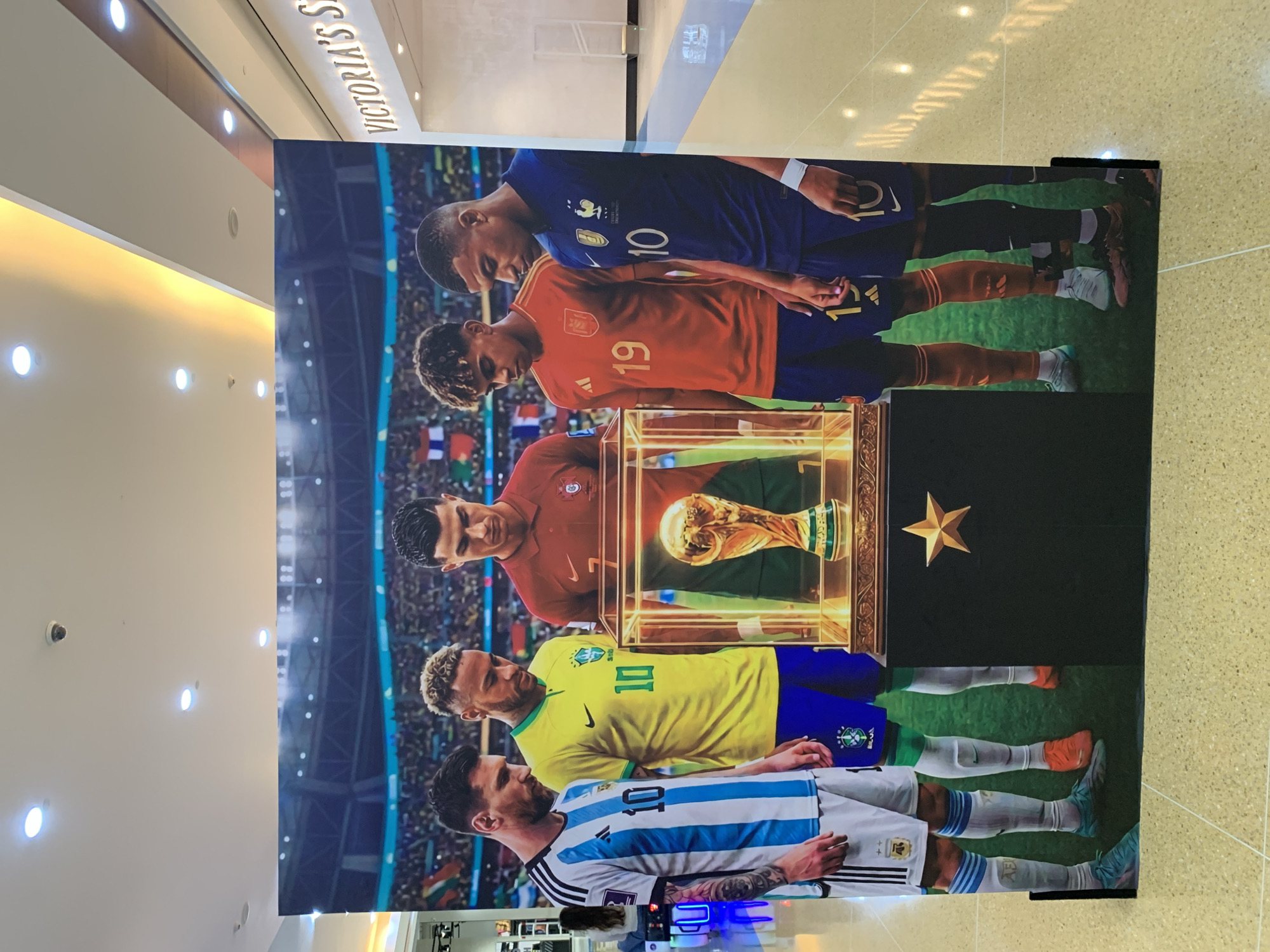 The image shows a large, freestanding cutout of five famous soccer players standing around a golden trophy on a pedestal. From left to right, the players are Lionel Messi (Argentina), Neymar (Brazil), Cristiano Ronaldo (Portugal), and two unidentified players wearing Spain and France national team jerseys. The backdrop depicts a soccer stadium crowd. The cutout is positioned in a brightly lit indoor space, likely a shopping mall, with a "VICTORIA'S SEcret" sign visible in the background on the right. The floor is tiled and reflective. No specific details about Porlamar, Venezuela are present in the image itself.