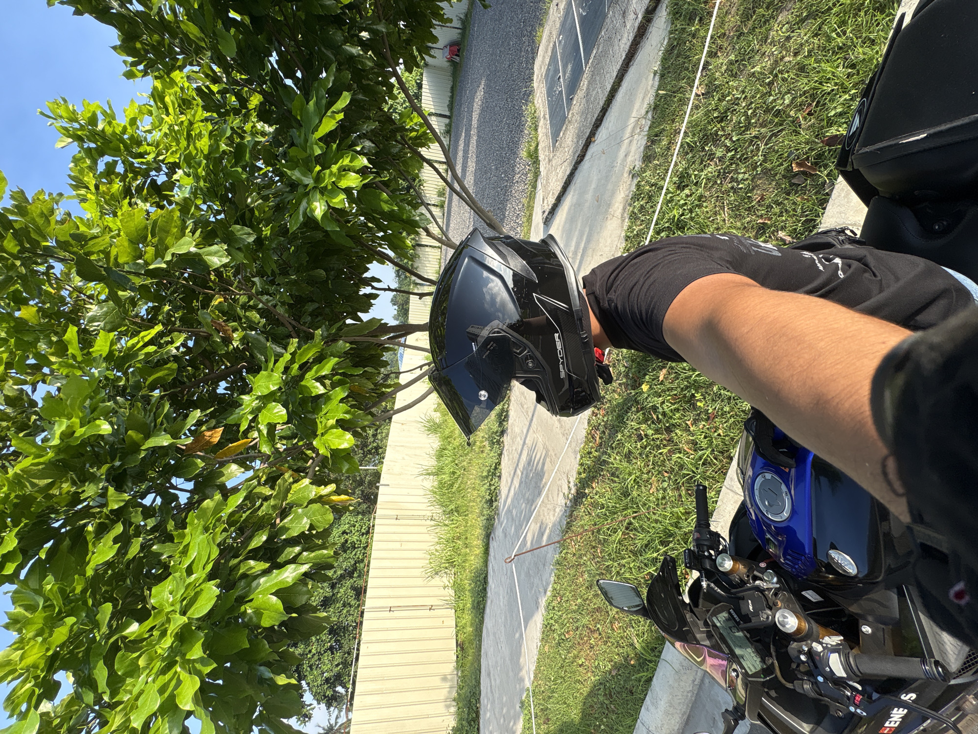The image shows a first-person perspective of someone sitting on a motorcycle, wearing a black helmet. The motorcycle has a blue and black fuel tank and visible parts of the handlebars and dashboard. In the background, there is a light-colored corrugated metal fence, a gravel-covered area, and a large tree with green leaves. The setting appears to be outdoors, possibly in a parking lot or a yard. There is a white rope cordon across a concrete path in the foreground. The time of day appears to be daytime, with sunlight casting shadows. The location is identified as Davao City, Philippines, based on the provided information.