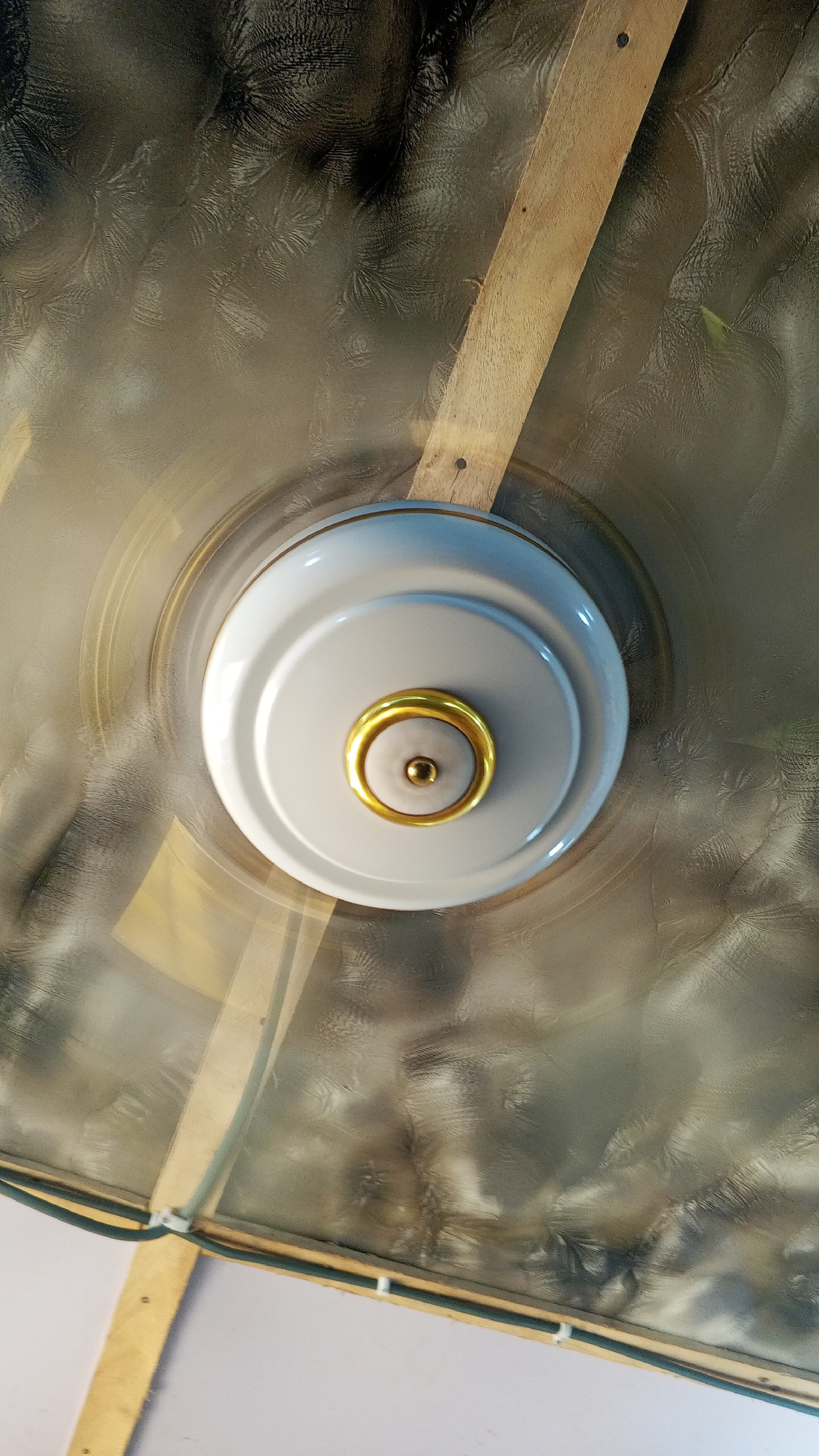 The image presents a direct overhead view of a ceiling fan in motion. The fan's central housing is white with prominent gold trim around its circular base and a central gold knob. Due to the fan's operation, its blades are blurred, creating translucent, swirling patterns that suggest rapid movement around the fixed white hub.
The ceiling itself is covered with a textured material, displaying intricate, dark, abstract patterns reminiscent of frost on a window or organic lace, set against a lighter, somewhat reflective background. Two wooden planks or beams are visible, traversing the ceiling diagonally. These wooden elements are light brown, showing wood grain, and are secured with visible nails. In the lower left section of the image, a dark green cable or wiring runs along one of the wooden planks. The setting is indoors. There are no people visible, nor are there any indications of the time of day, weather, or discernible emotions. No text is present in the image.