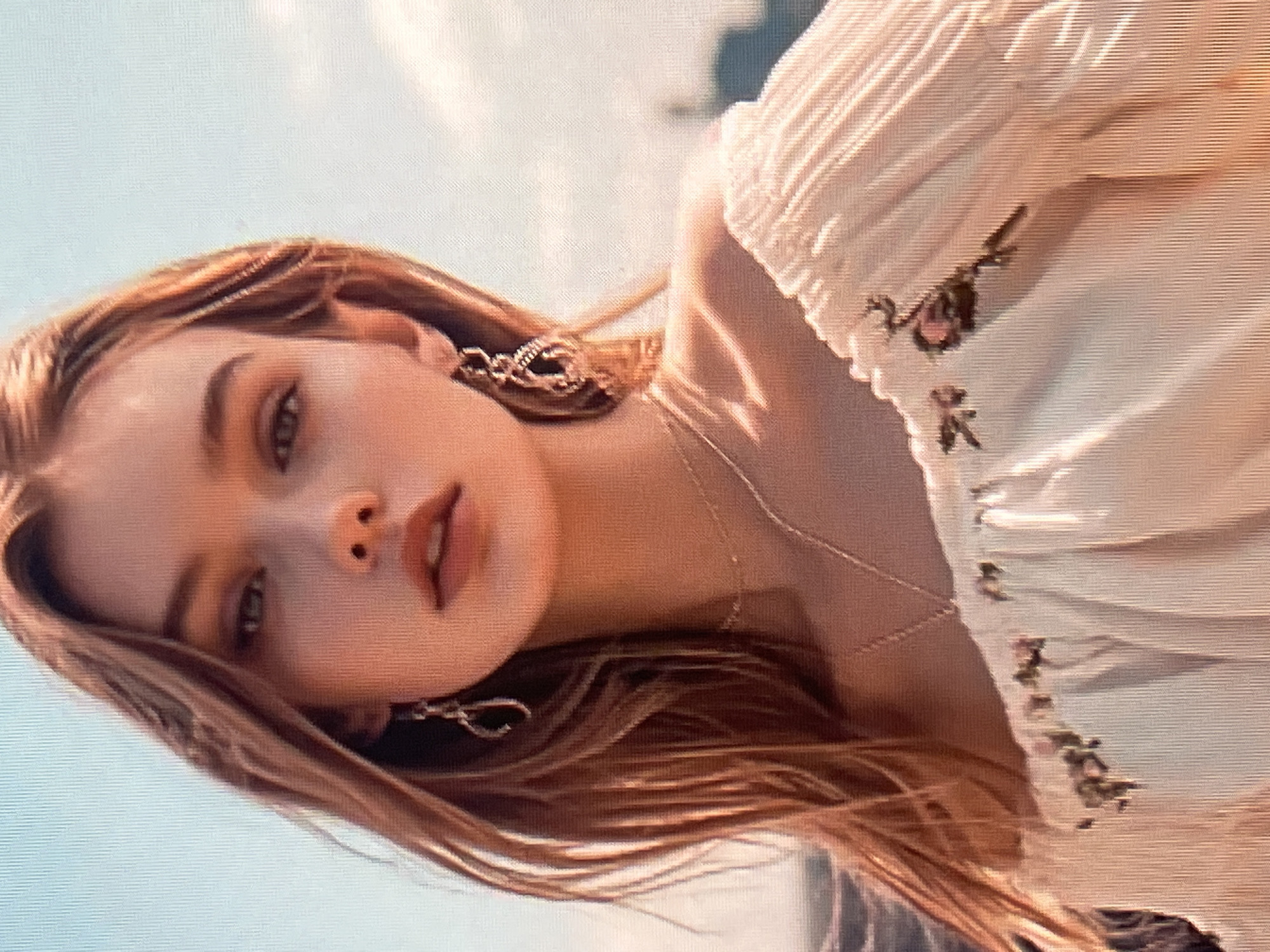 A close-up, waist-up shot shows a young woman with long, light brown hair and fair skin. She is wearing an off-the-shoulder white blouse with a ruffled neckline and embroidered floral details. She has on delicate necklaces and ornate dangling earrings. The background is a blurred sky with some distant structures visible, suggesting an outdoor setting in an urban environment. The lighting is warm and natural, possibly indicating daytime. There is no specific indication of the location being Qarağandı city administration, Kazakhstan.