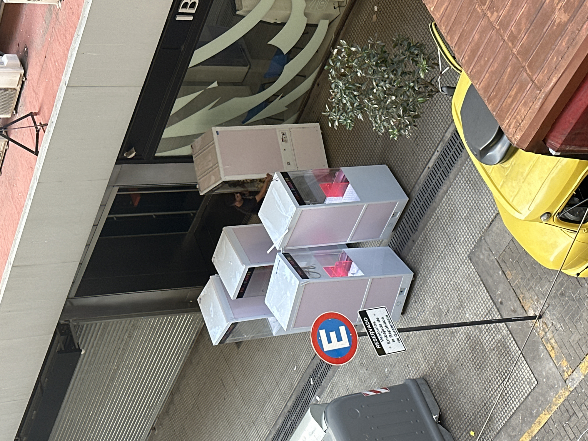 The image depicts a street scene in Buenos Aires, Argentina. Three white, cube-shaped structures are stacked vertically near a building's facade. The structures appear to be advertising kiosks or display cases, with transparent tops and interior lighting illuminating red elements inside. A "No Parking" sign with a blue "E" in a red circle is visible, along with a smaller sign indicating the parking space is reserved for vehicles of the Embassy of Guatemala. A section of a yellow car is parked on the right side of the frame. A small tree stands near the car. A grey dumpster is visible near the "No Parking" sign.