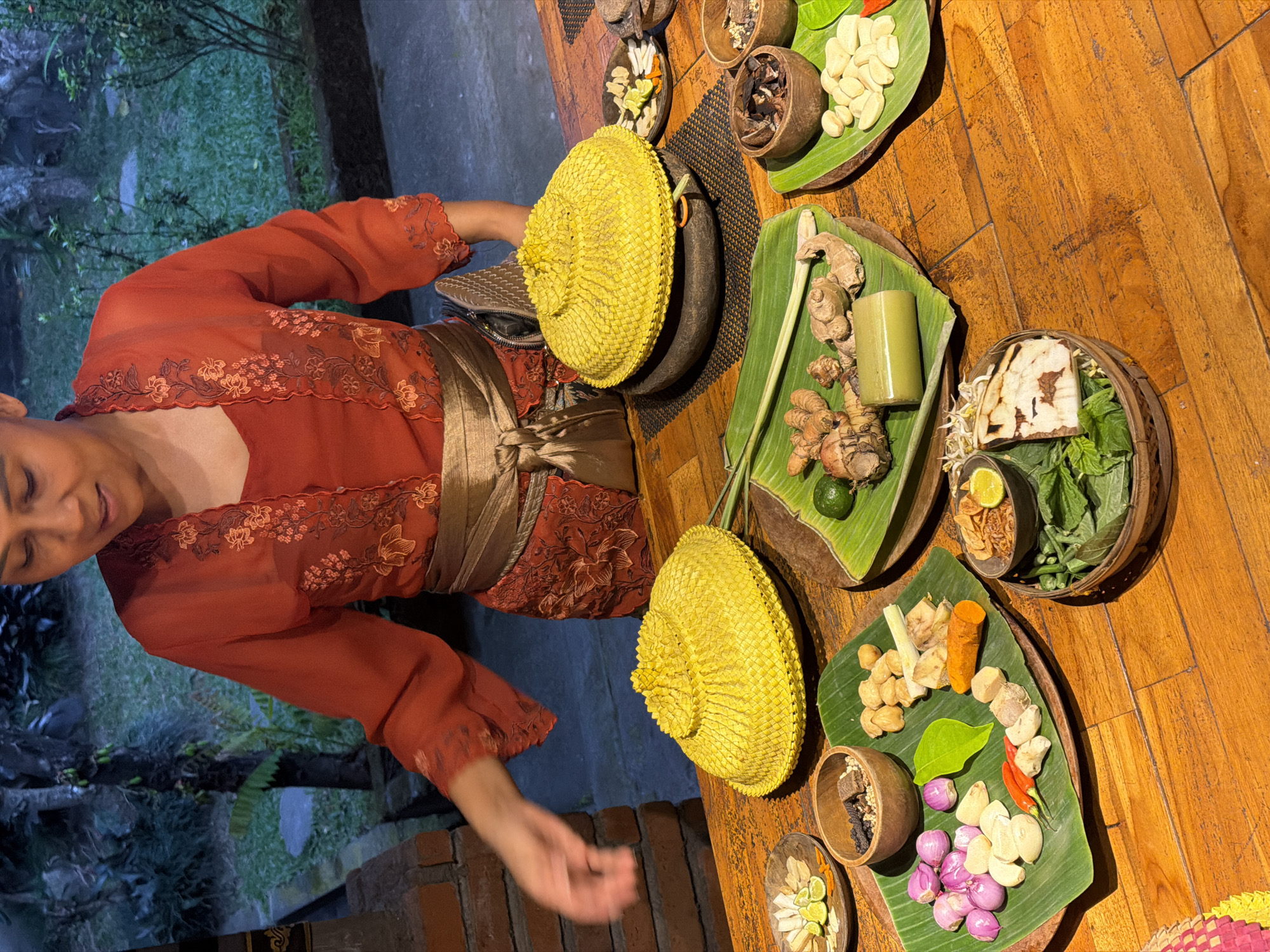 In Mengwi, Indonesia, a woman dressed in a traditional orange outfit with gold trim stands behind a wooden table displaying various ingredients for a culinary preparation. The ingredients, arranged on banana leaves in small wooden bowls and on plates, include shallots, garlic, chili peppers, ginger, turmeric, lemongrass, nuts, dried herbs, and sliced limes. Two large, woven yellow lids, possibly for steaming or cooking, are prominently placed on the table. The background suggests an outdoor setting with greenery and trees visible. The woman's hand is extended forward, indicating she may be in the process of preparing or presenting the ingredients.