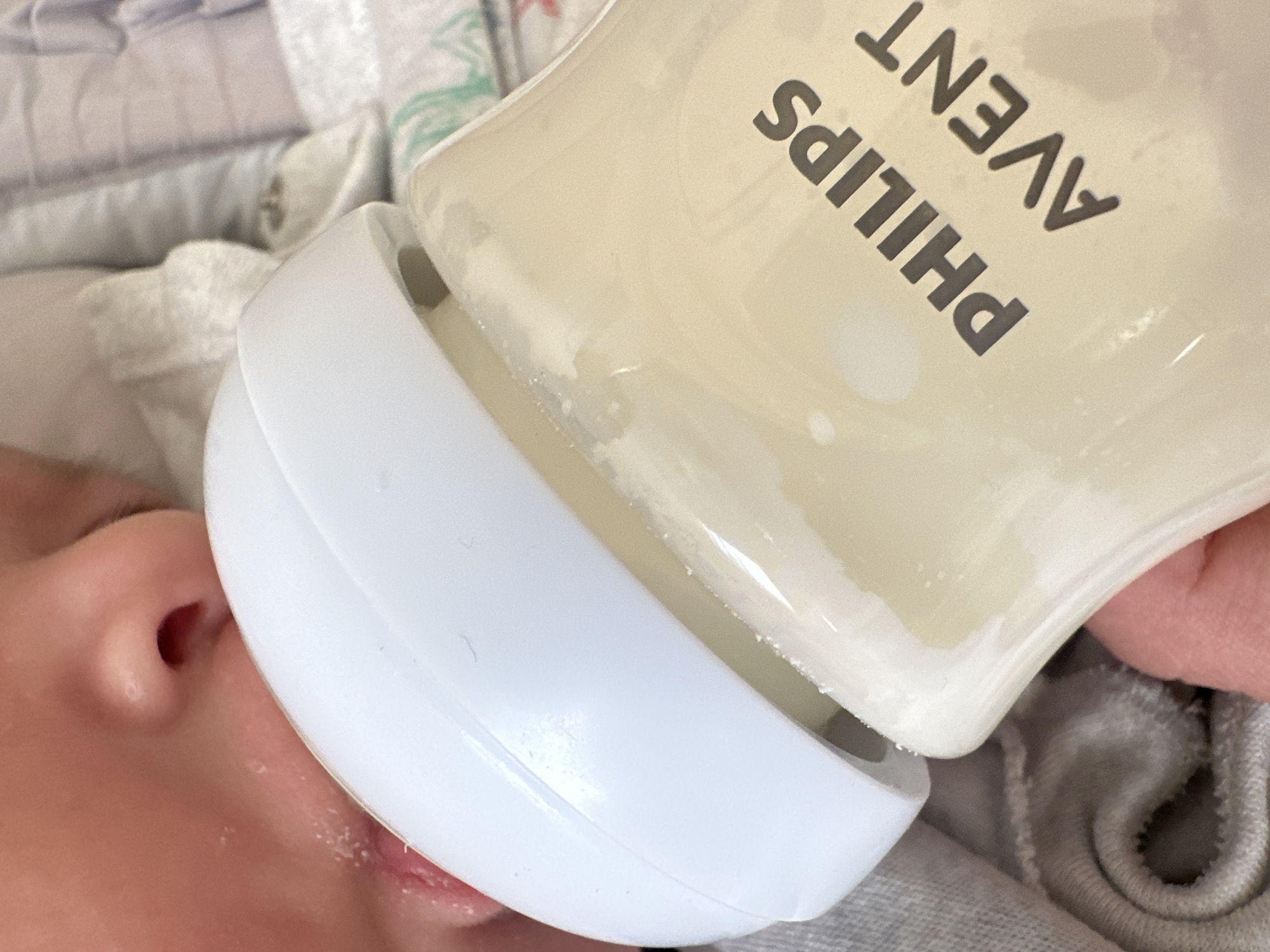 A close-up, eye-level shot shows a baby's mouth and nose as they are feeding from a Philips Avent baby bottle filled with a light-colored liquid. The baby's lips are slightly parted, and their nose is visible above the white bottle nipple. The bottle is clear and shows the liquid level. The branding "PHILIPS AVENT" is visible in dark gray lettering on the side of the bottle. The background consists of soft, textured fabric, possibly a blanket or clothing, in muted tones of gray and light blue, with some green and white patterns. The lighting is soft and diffused, highlighting the textures and colors of the subjects. The scene appears to be indoors, in a domestic setting. There is no identifiable location in Masazir, Azerbaijan, present in this image.