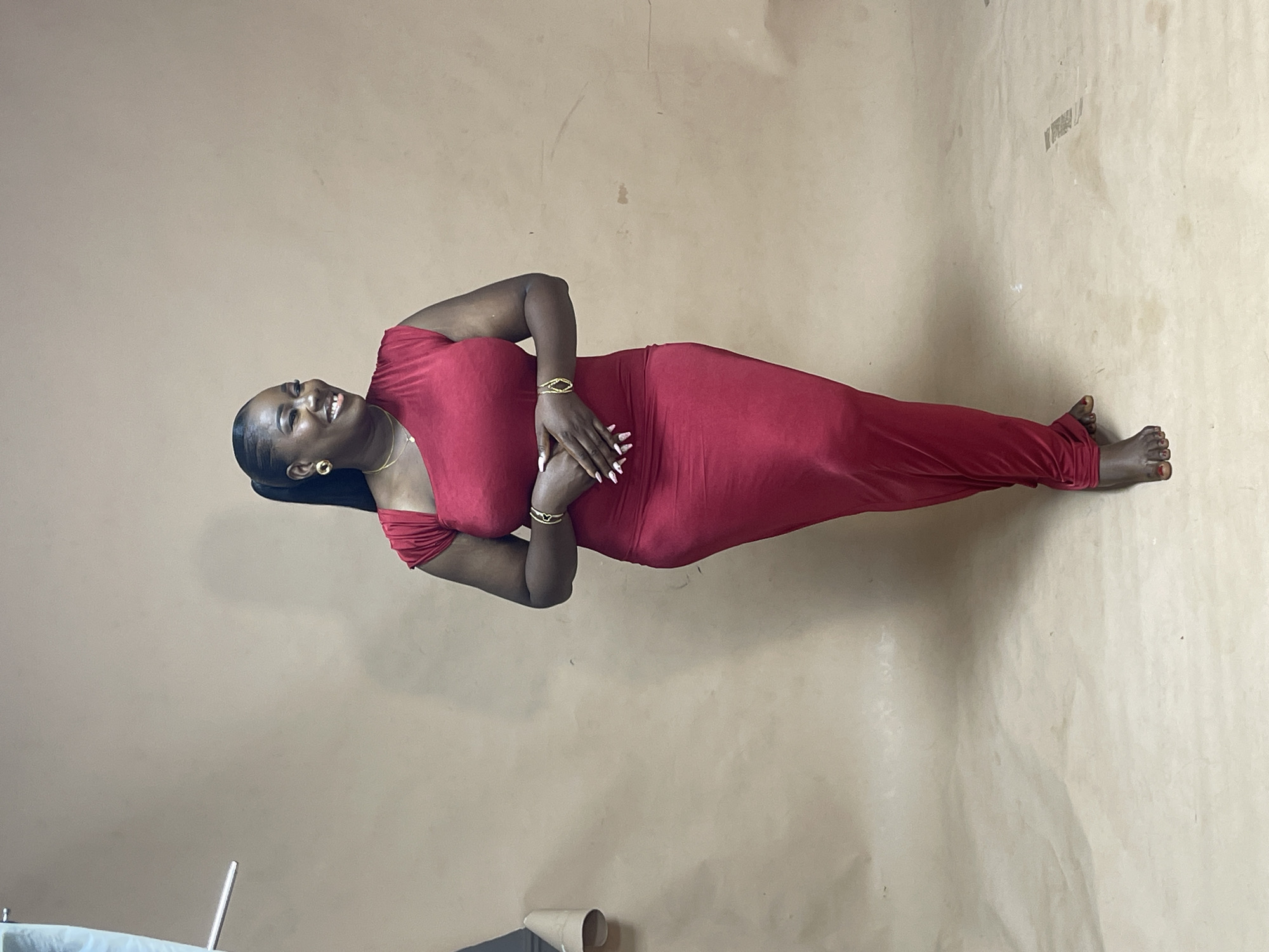 A dark-skinned woman wearing a form-fitting red dress poses against a tan backdrop. She is smiling and has her hands clasped near her abdomen. She is wearing gold bracelets and earrings. The backdrop appears to be a sheet of paper, potentially used as an impromptu photo background. The location is not evident from the image. It is not possible to determine if the location is in Abuja, Nigeria, or any other place.