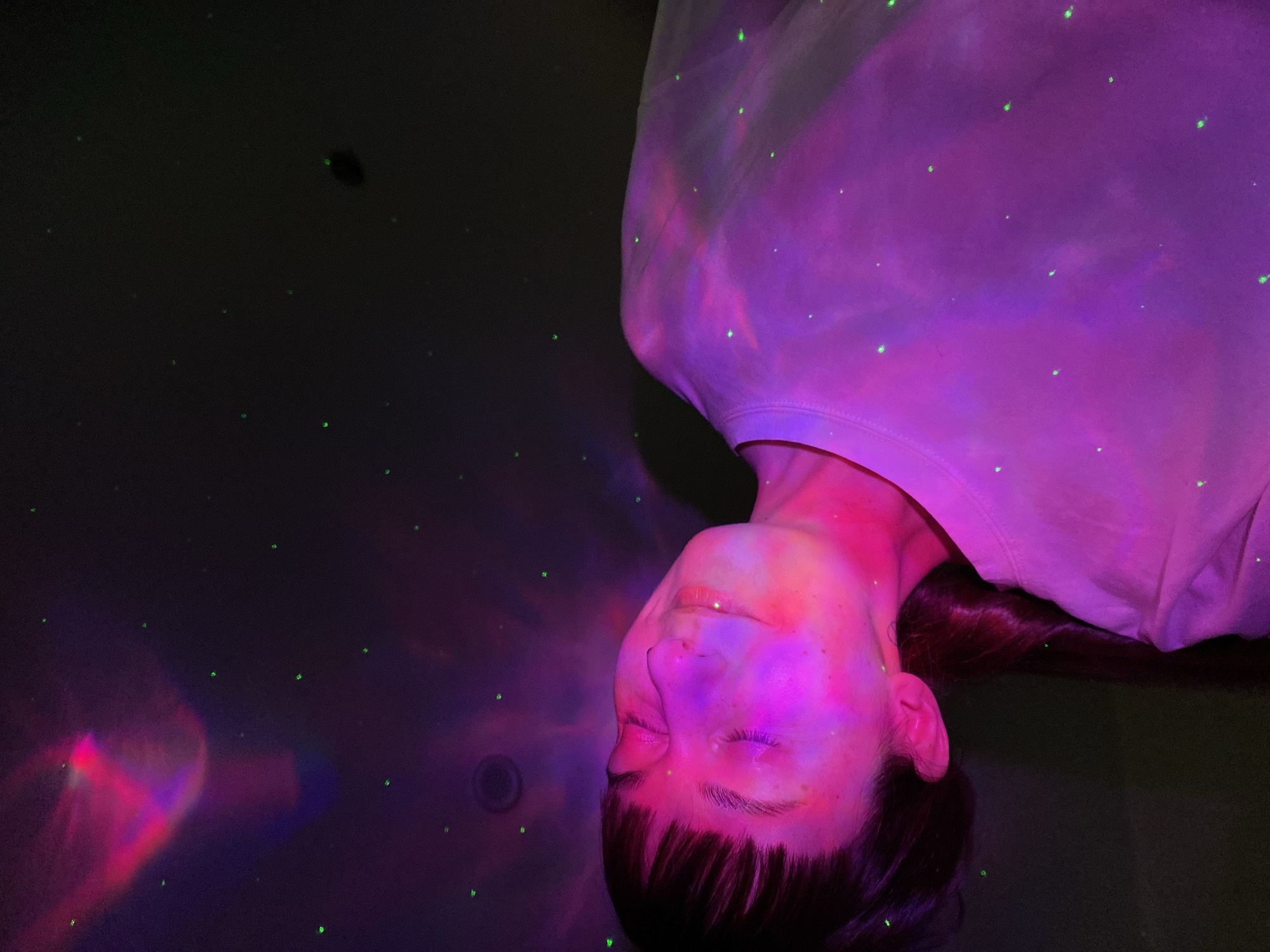 A woman lies supine on a dark surface. Her face, illuminated with pink and purple light, is centered with her head near the bottom of the frame. She has dark hair, fair skin, and her eyes are closed. A white t-shirt covers her upper torso. Green dots are scattered across the scene, resembling stars. To the left of the woman, a swirl of red and purple light creates a hazy, nebula-like effect. The location is unknown, but the prompt suggests Votkinsk, Russia. The scene implies a relaxed state in a room with colored lighting.