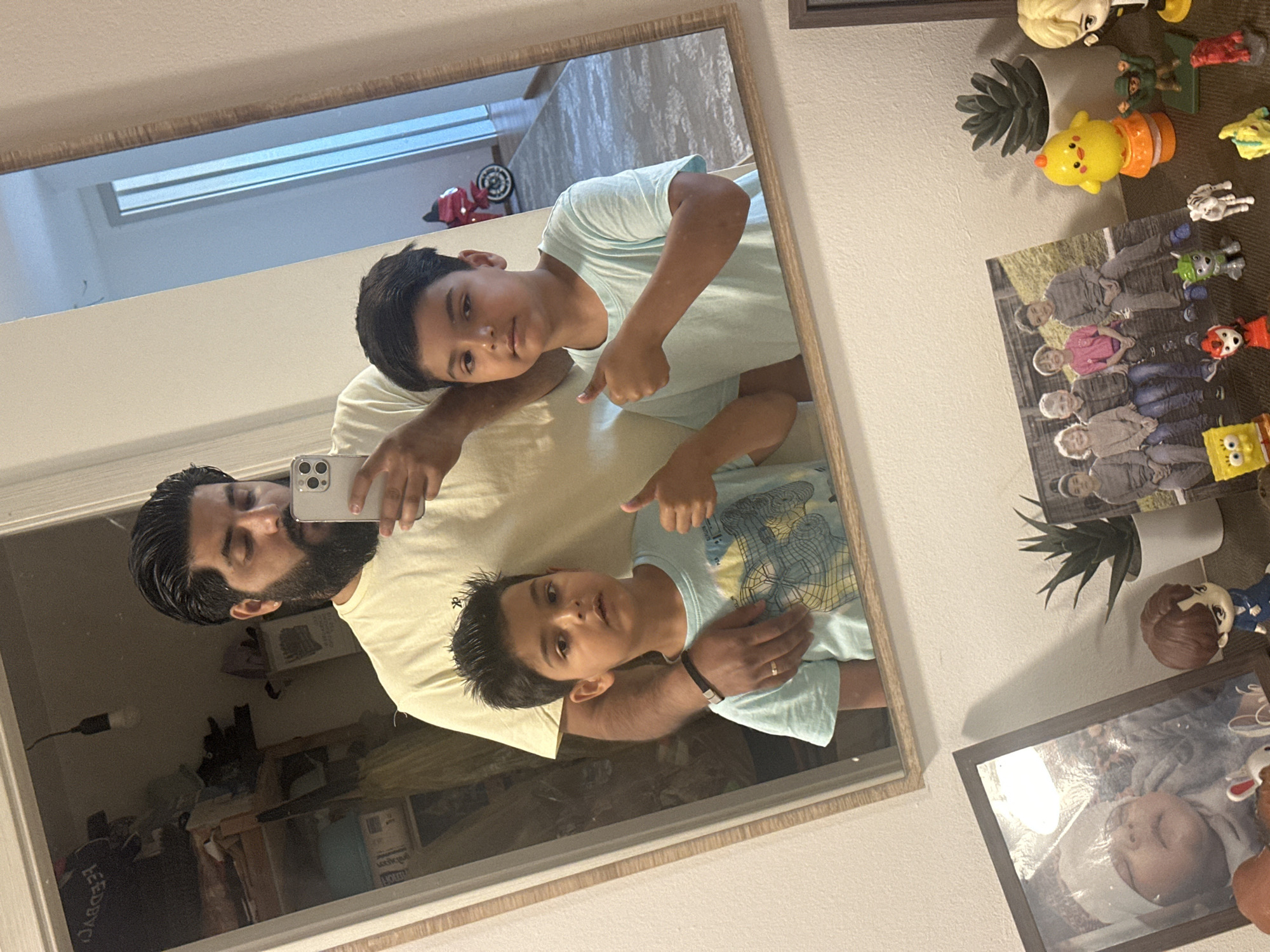 A man is taking a mirror selfie with two young boys in what appears to be a residential interior. The man, with dark hair and a beard, is wearing a light yellow t-shirt and holding a smartphone. The two boys, also with dark hair, are standing in front of him. The boy on the left is wearing a light blue t-shirt with a graphic print and is giving a thumbs-up. The boy on the right is wearing a light blue t-shirt and is also giving a thumbs-up. The background reflected in the mirror shows a doorway, a red toy motorcycle, and a hallway. On a shelf below the mirror, there are several framed photographs and small figurines, including a framed picture of a baby, a group photo of children, and various cartoon character figurines. The overall environment suggests a home setting, possibly in Mainburg, Germany, given the context provided.