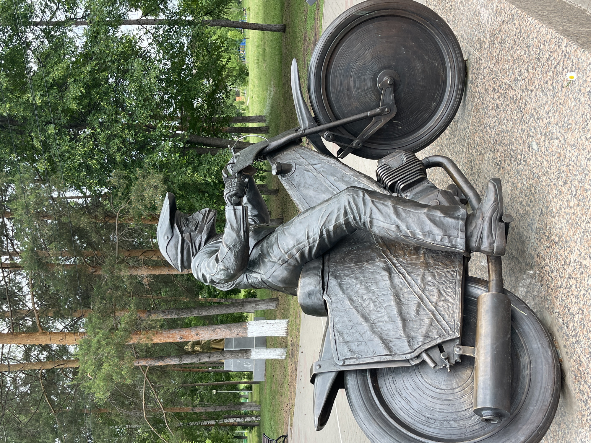 A bronze statue of a motorcyclist is prominently featured in an outdoor park setting, located in Oktyabrsky, Russia. The statue depicts a single figure seated on a detailed motorcycle, both rendered in a dark, metallic bronze finish. The motorcyclist wears a full-face helmet, a textured jacket, and riding trousers, with hands gripping the handlebars and feet positioned on footpegs, conveying a dynamic riding posture. The motorcycle itself includes intricate details such as spoked wheels (though appearing solid in the bronze), an engine with cooling fins, an exhaust pipe, and a sculpted seat.
The background consists of a lush green park or forest area. Tall pine trees with reddish-brown trunks and green foliage are visible, alongside other deciduous trees, suggesting a diverse arboreal landscape. A paved pathway runs through the park, and patches of green grass are visible. In the middle distance, faint white rectangular structures or monuments can be discerned among the trees. The lighting suggests it is daytime, with soft, diffused light, indicating either an overcast sky or dappled sunlight filtered through the trees. No visible text is present on the statue or in the immediate surroundings. The overall scene captures a moment of potential motion and the enduring spirit of motorcycling within a natural, public space.