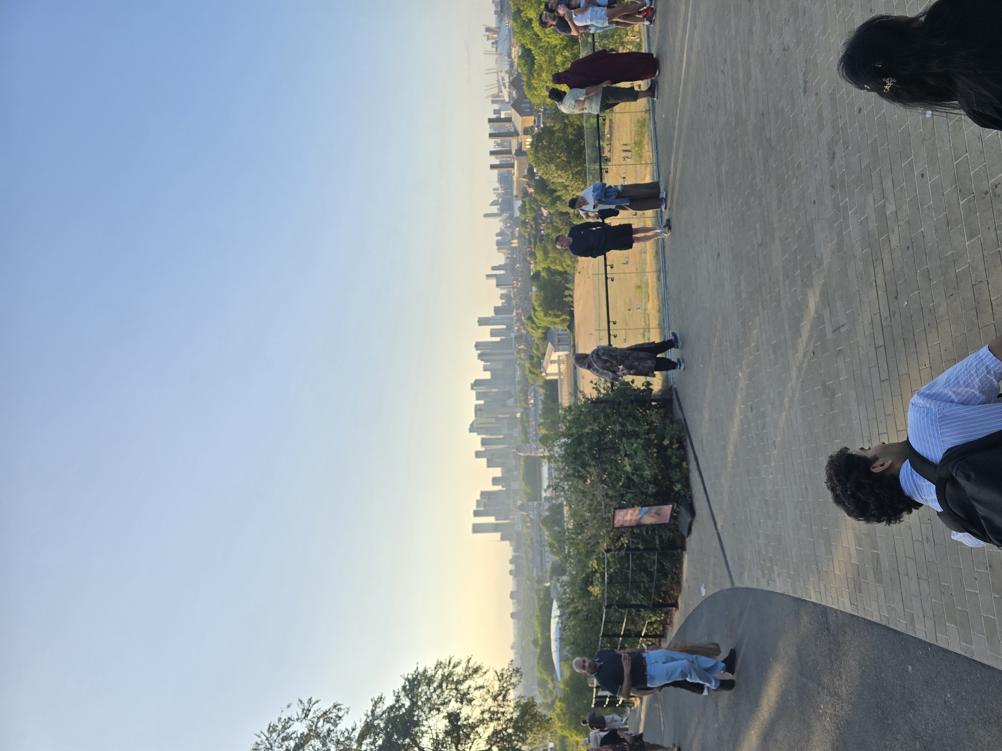 The image depicts a view of the London skyline from Greenwich Park, United Kingdom. In the foreground, a paved pathway curves to the left, with people walking and standing along a low railing. A man in a blue and white striped shirt and a black backpack is seated in the lower right corner, facing away from the camera. Further along the pathway, a group of people are gathered, some observing the view. To the left, a man in light blue jeans and a dark polo shirt walks down the path. The middle ground features lush green trees and a large open field. In the background, the iconic London skyline is visible, characterized by numerous skyscrapers and other urban structures. The sky above is a clear, pale blue, suggesting a bright day.