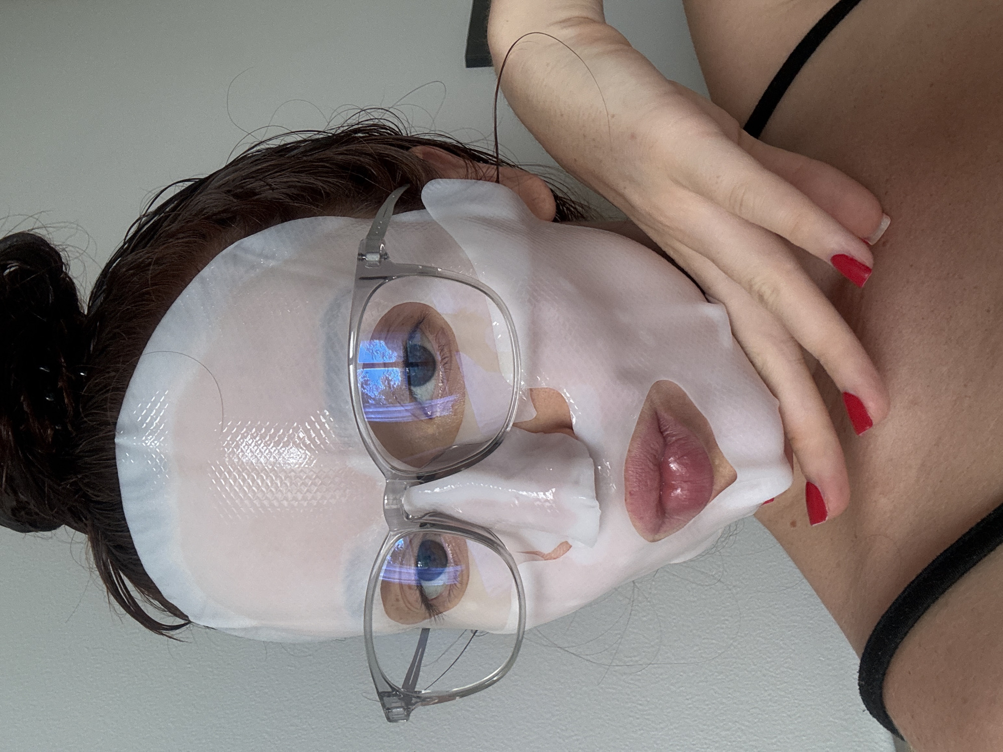 A woman with brown hair pulled back in a bun is wearing a white sheet mask and clear-framed eyeglasses. The mask covers most of her face, leaving openings for her eyes and mouth. She has red nail polish on her fingernails and is touching her neck with her hand. The scene is indoors, with a white wall visible. No specific location in Choeng Thale, Thailand, is evident in the image.