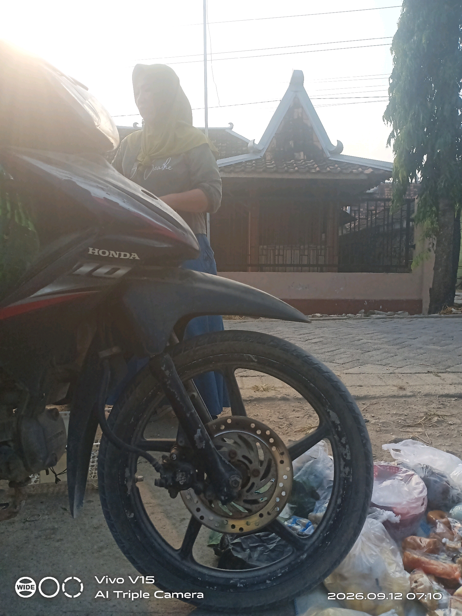 In Plosorejo, Indonesia, a person wearing a light yellow hijab and a grey long-sleeved shirt is standing next to a black motorcycle. The motorcycle has a "HONDA" logo on its side. The scene appears to be outdoors, possibly near a building with a distinctive tiled roof and a fence. To the right of the motorcycle, there is a pile of plastic bags containing various items. The ground is paved with bricks. A date and time stamp in the lower right corner indicates "2026.09.12 07:10".