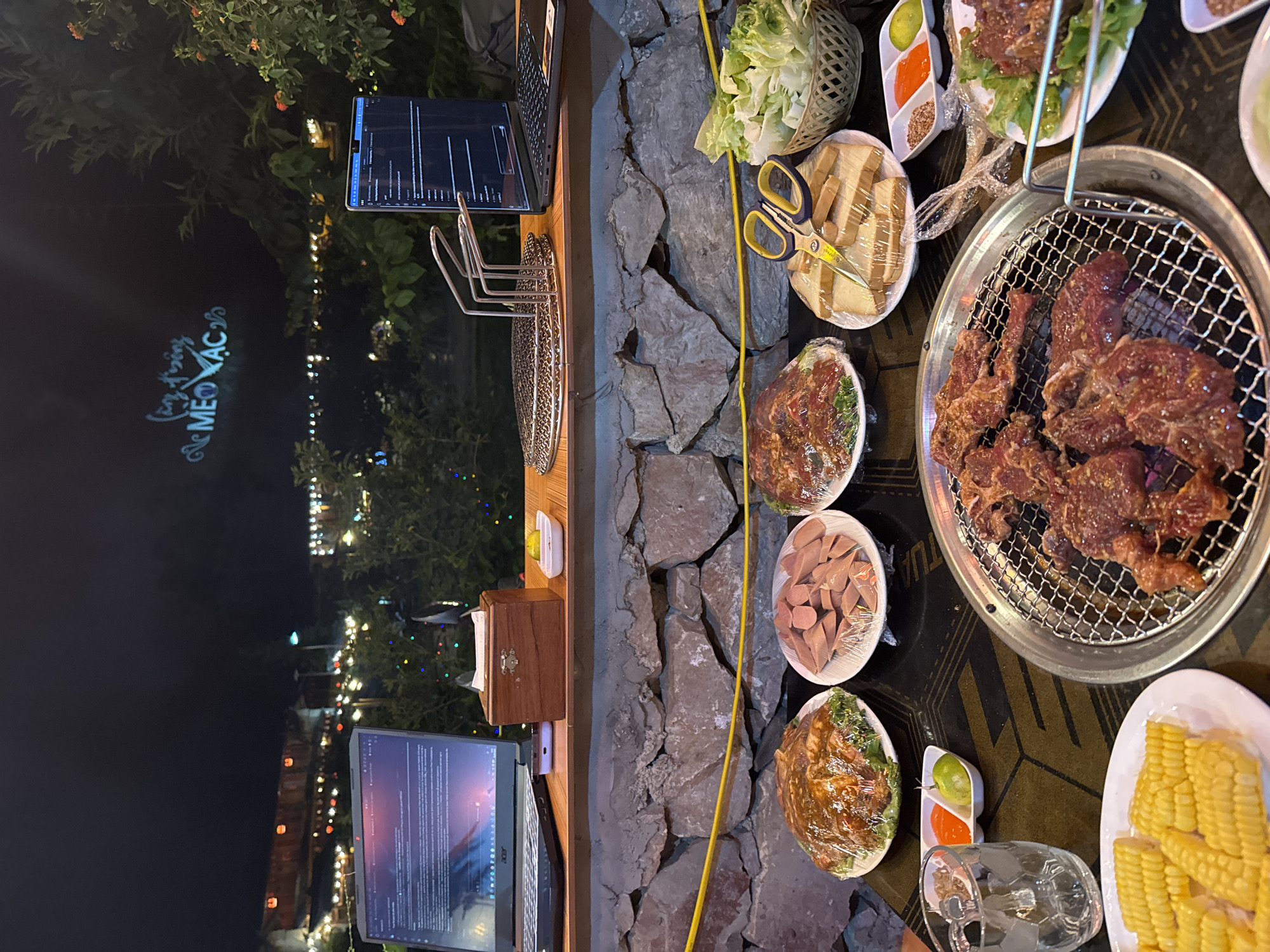The image shows a barbecue setup, likely in Mèo Vạc Commune, Vietnam, based on the illuminated sign in the background. The scene is outdoors at night. The barbecue is centered around a round grill with meat cooking on it. Adjacent to the grill are plates with various food items, including lettuce, sausages, bread, marinated meat, corn on the cob, and condiments. A pair of scissors is placed near the bread. A table extends to the left of the barbecue, holding a laptop displaying what appears to be a code editor and a smartphone showing charts. A wooden box and a grill rack are also on the table. Street lights and foliage are visible in the background.