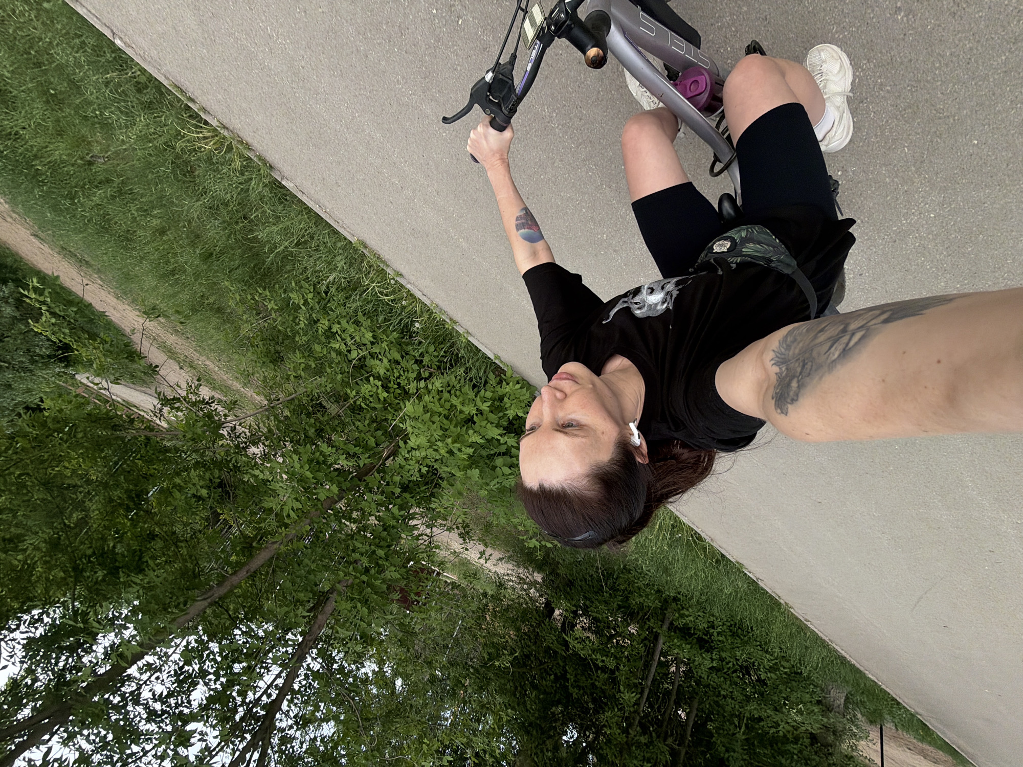 A woman is taking a selfie while riding an electric bicycle along a paved path. She is wearing black shorts, a black t-shirt, and white sneakers. A patterned waist bag is around her waist. She has a tattoo on her forearm and is wearing an earpiece. The bicycle is silver, and a purple water bottle holder is attached to the frame. The path is bordered by green grass and trees. The location is the city of Perm, Russia.