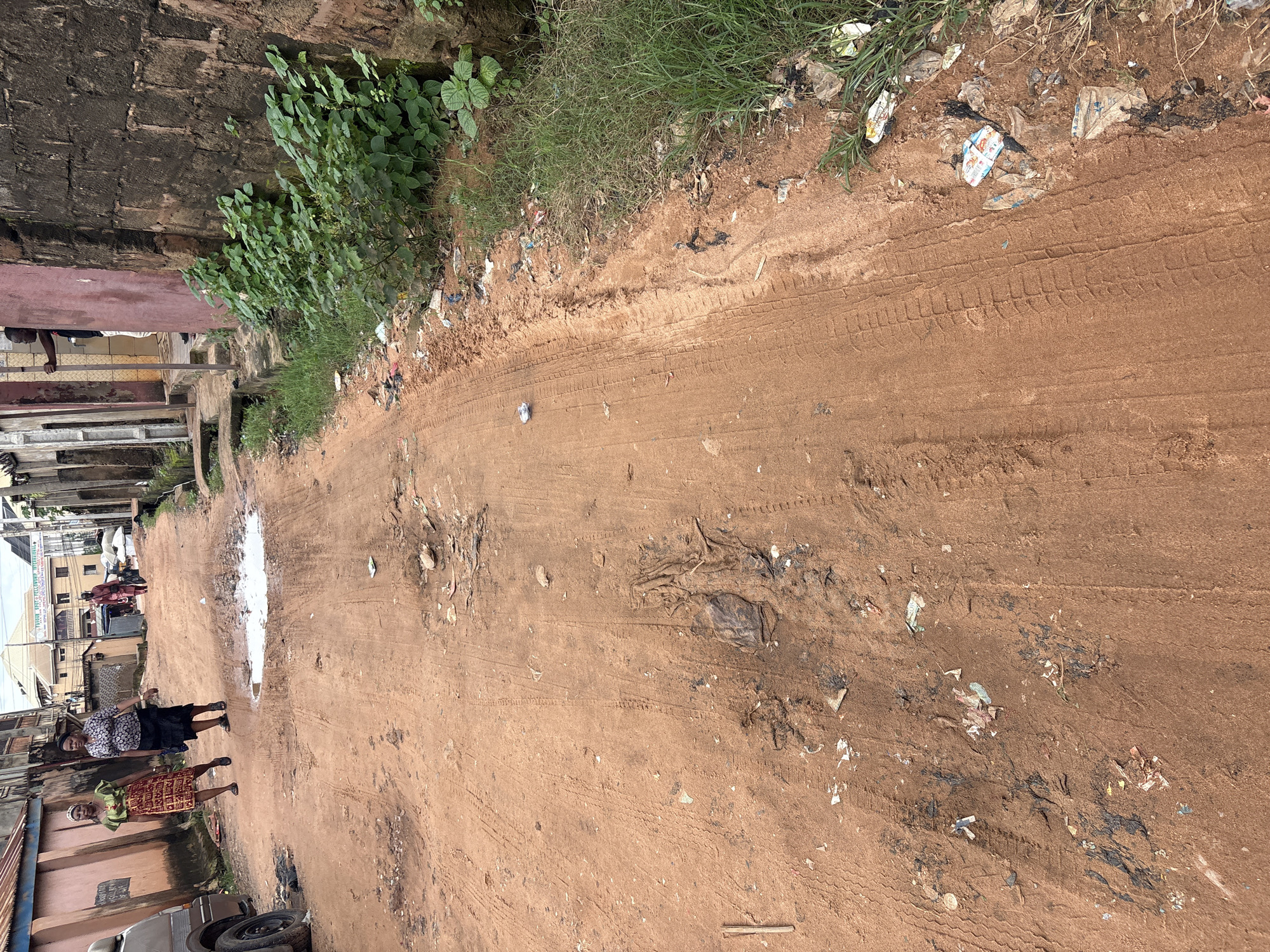 In Nkpor, Nigeria, a dusty, reddish-brown dirt road is littered with debris and tire tracks. Two women walk on the road, one in a green and yellow patterned dress and the other in a black and white patterned top and black skirt. In the background, a person in a red garment walks away from the camera, and further down the road, buildings line the street. Green foliage grows along the right side of the road, interspersed with more trash. A small puddle of water is visible in the road ahead of the women.