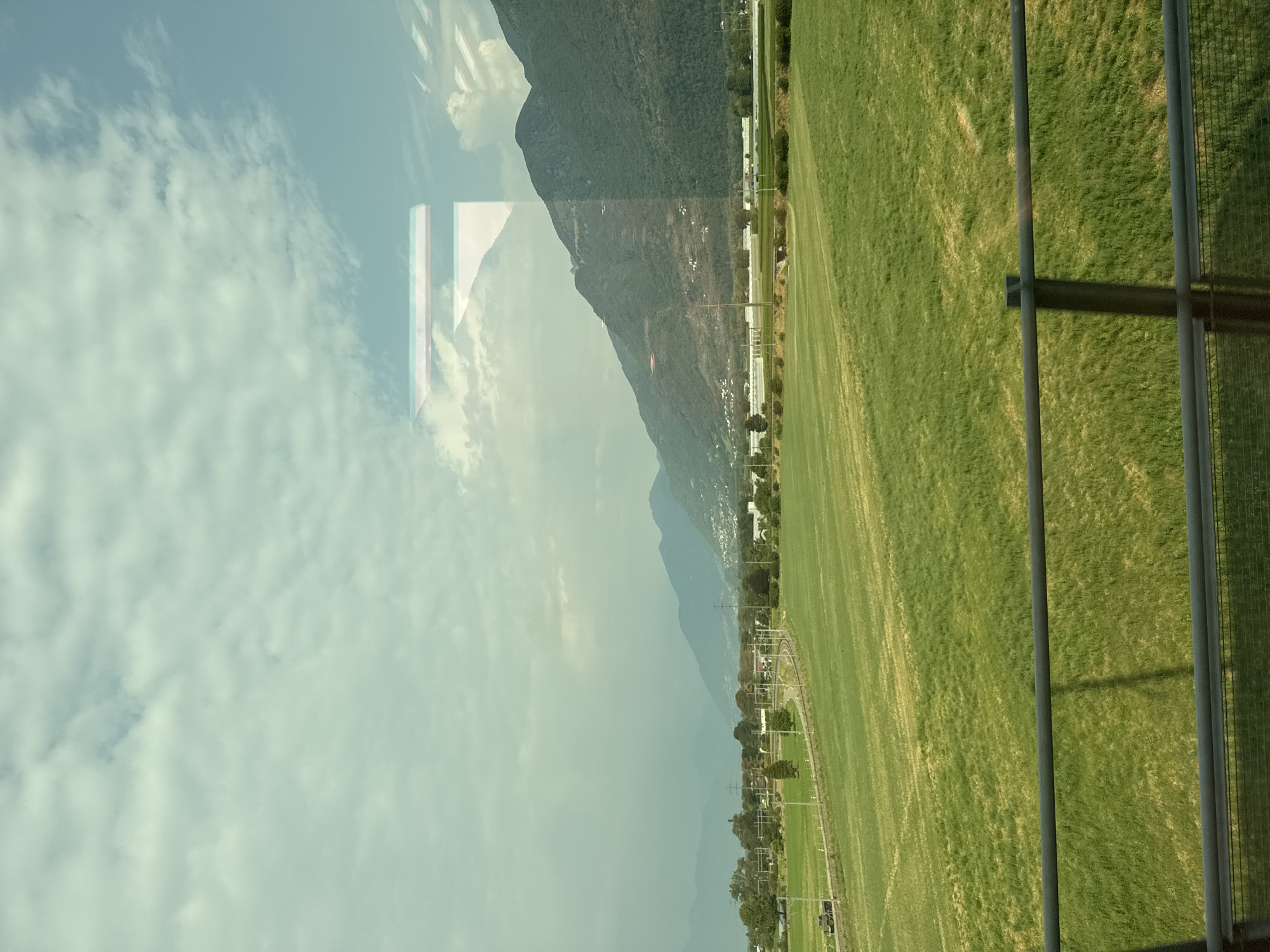 The image captures a landscape view, likely from a moving vehicle, showcasing a vast expanse of green, undulating fields. In the mid-ground, a railway track curves through the fields, with a dark-colored train visible on it. Scattered trees dot the landscape, and a few buildings are nestled in the distance. Beyond the fields and buildings, a range of mountains forms the backdrop, with some of the slopes appearing cultivated or dotted with structures. The sky is partly cloudy, with a hazy quality that suggests either distance or atmospheric conditions. Reflections on what appears to be a window are visible in the upper portion of the image, creating abstract shapes and obscuring parts of the sky and mountains. The scene is predominantly green and blue, with muted tones.