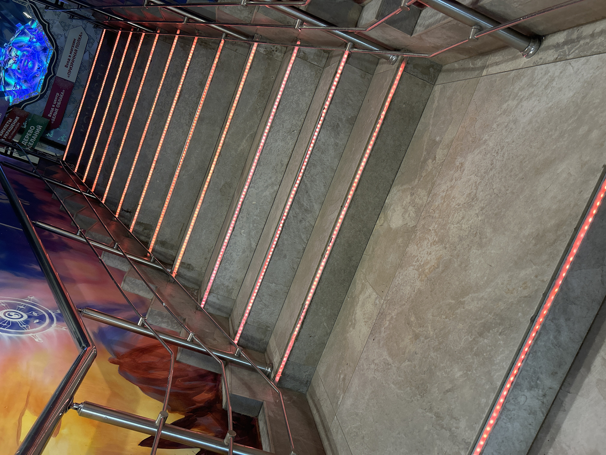 A high-angle shot captures a stairwell in Unknown, Unknown. The stairs are concrete with linear red LED lighting installed in the risers. Stainless steel handrails run along the sides. On the left, the wall is decorated with a fantasy-themed mural, including a large wheel and abstract landscape. Signage in Russian is visible on the upper left, indicating attractions like "White Clouds" and "Ghost Shelf," also referencing amulets, ornaments, and a "Wishing Tree." The right wall has a beige stone tile finish.