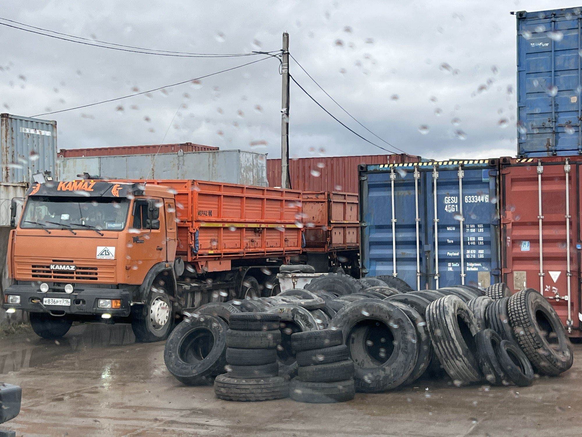 The image shows an orange KAMAZ truck parked in what appears to be a depot or storage area in Naberezhnye Chelny, Russia. The truck has a flatbed and the license plate "K836PT 716". The "KAMAZ" name is prominently displayed above the windshield. In front of the truck is a large pile of used tires. Behind the truck and tires are several shipping containers stacked on top of each other, some blue and some red. A utility pole with wires is visible in the background. The ground is wet, suggesting it has been raining. The sky is overcast. The image was likely taken through a wet windshield, as evidenced by the water droplets on the glass. The location is suggested by the KAMAZ truck brand (a Russian manufacturer), license plate number of Russia and the "MEFAZ" inscription on the truck (a possible reference to a regional business or organization).
