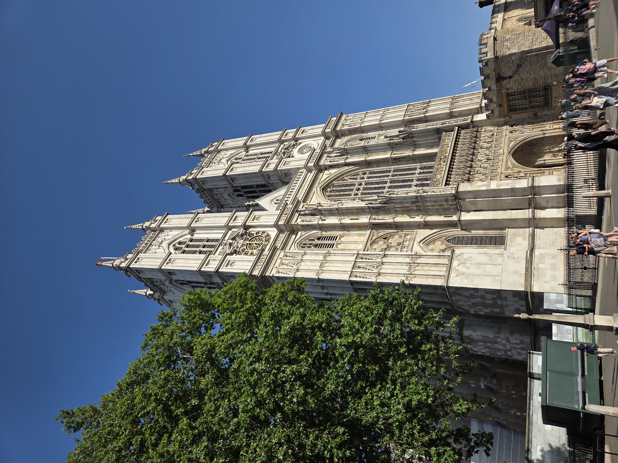 The image shows a partial view of Westminster Abbey in London, United Kingdom. The abbey is depicted from a low angle, showcasing its western facade, including its two towers, intricate stone carvings, and large windows. The facade is off-white or light gray, contrasting with the clear blue sky. A large, leafy green tree partially obscures the lower portion of the abbey. A wrought iron fence runs along the side of the abbey, separating it from a sidewalk. A small group of pedestrians are visible, walking along the sidewalk adjacent to the fence. A green booth or structure is also present along the sidewalk, near the abbey. The scene is bathed in daylight.