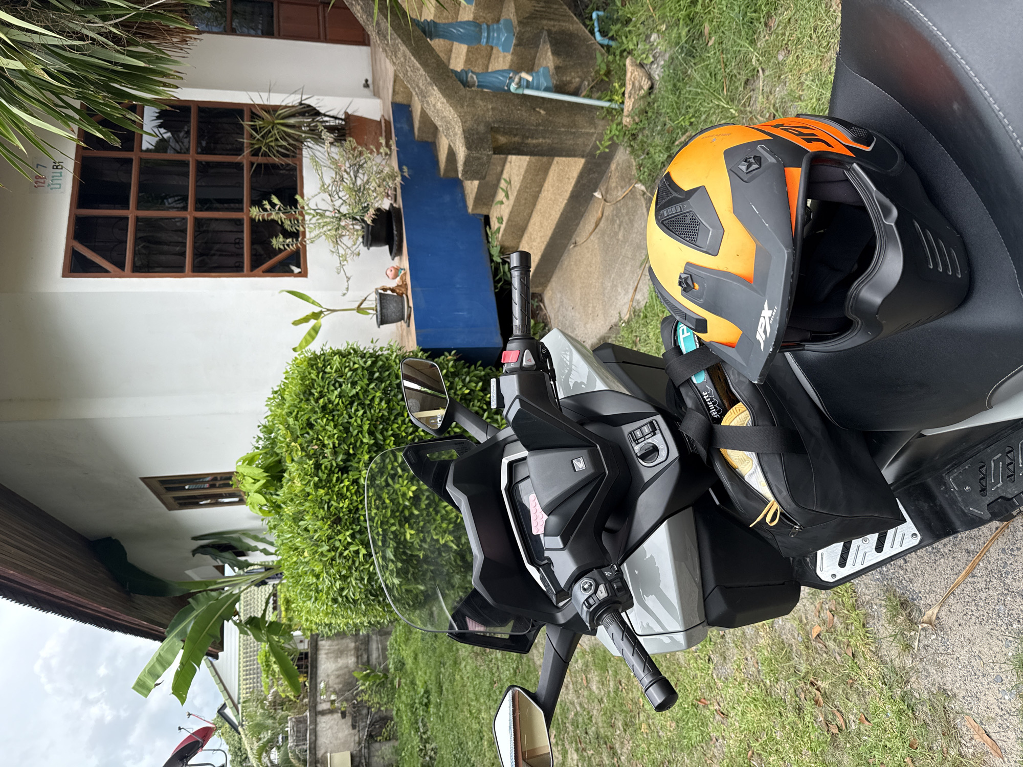 A grey scooter is parked on a gravel path in front of a white house with a wooden-framed window. An orange and black helmet rests on the seat of the scooter, partially obscuring a black bag. Lush green vegetation surrounds the scooter and the house. A portion of a concrete stairway with blue railings is visible to the right of the scooter. The sky is overcast. The location is identified as บ้านบางรักษ์, Thailand.