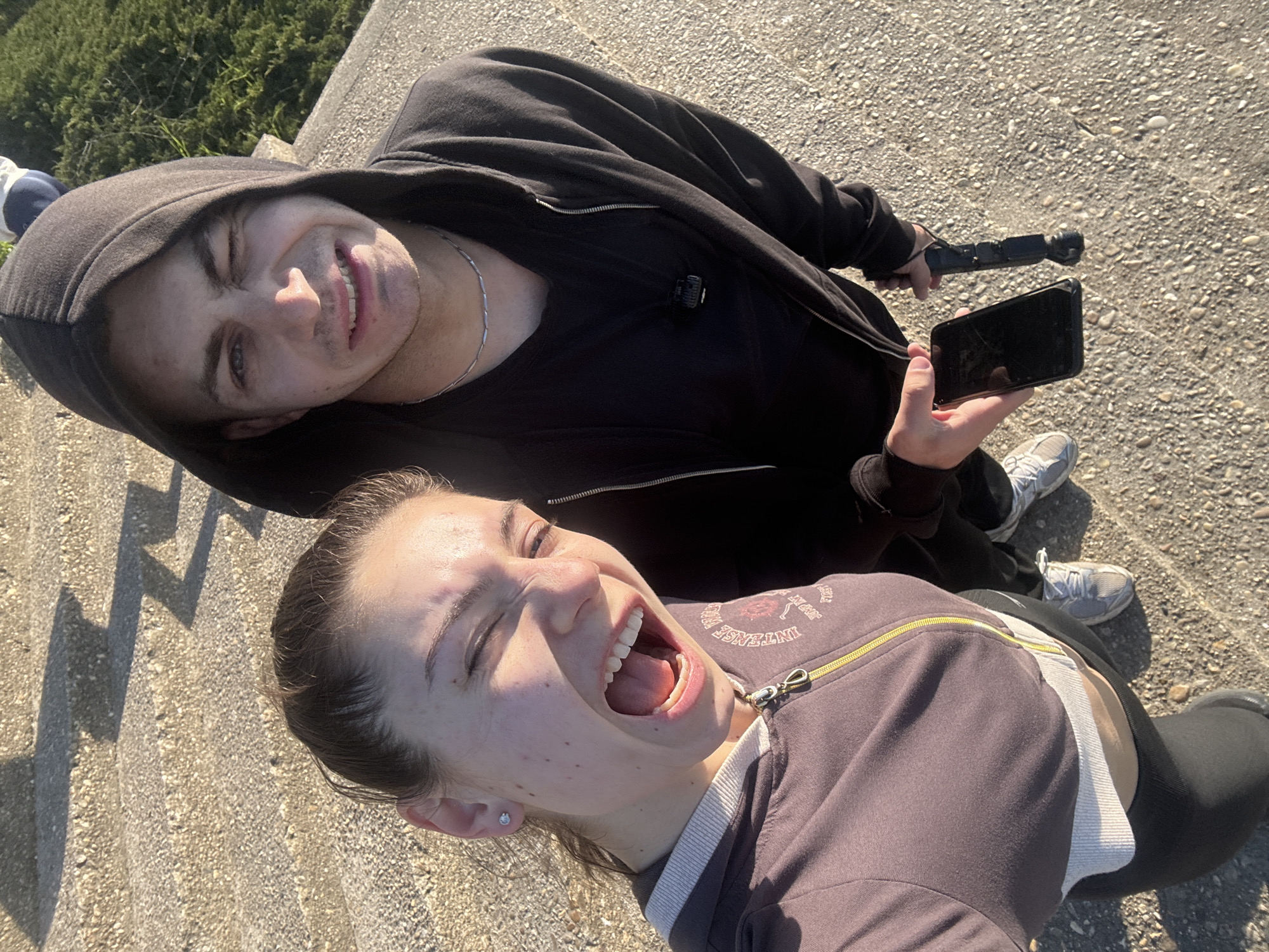 Two individuals, a male and a female, are captured in a selfie taken from an elevated position. The male, positioned above, wears a black hooded jacket over a black t-shirt, and a silver chain is visible around his neck. He is holding a black smartphone in his left hand and a black handheld device in his right hand. The female, positioned below, wears a dark brown jacket with a visible zipper. Both individuals are facing upwards towards the camera with expressive smiles. The background is a concrete surface under sunlight. The location is Varna, Bulgaria.