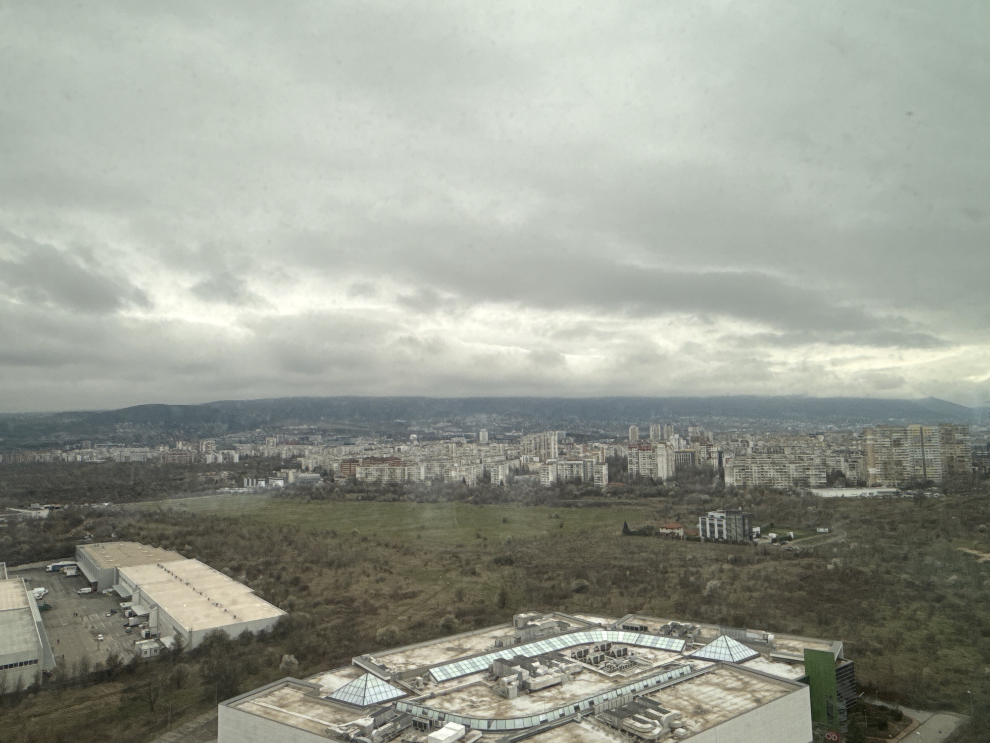 The image shows a city view from an elevated position, likely a high-rise building, on an overcast day. In the foreground, the rooftops of modern structures with glass and metal elements dominate the scene. Some are shopping malls with visible glass roofs and skylights. A large, flat-roofed warehouse-type building is seen to the left. Beyond these structures is a large green space, appearing to be a park or undeveloped area, bordered by bare trees. In the midground is the sprawling urban landscape of Sofia, Bulgaria, characterized by dense clusters of buildings, mostly residential, extending to the foothills of a distant mountain range. The sky is heavily overcast, obscuring details of the skyline and giving the entire scene a muted, grey tone. There are no visible people in the image. The location is in the city of Sofia, Bulgaria.