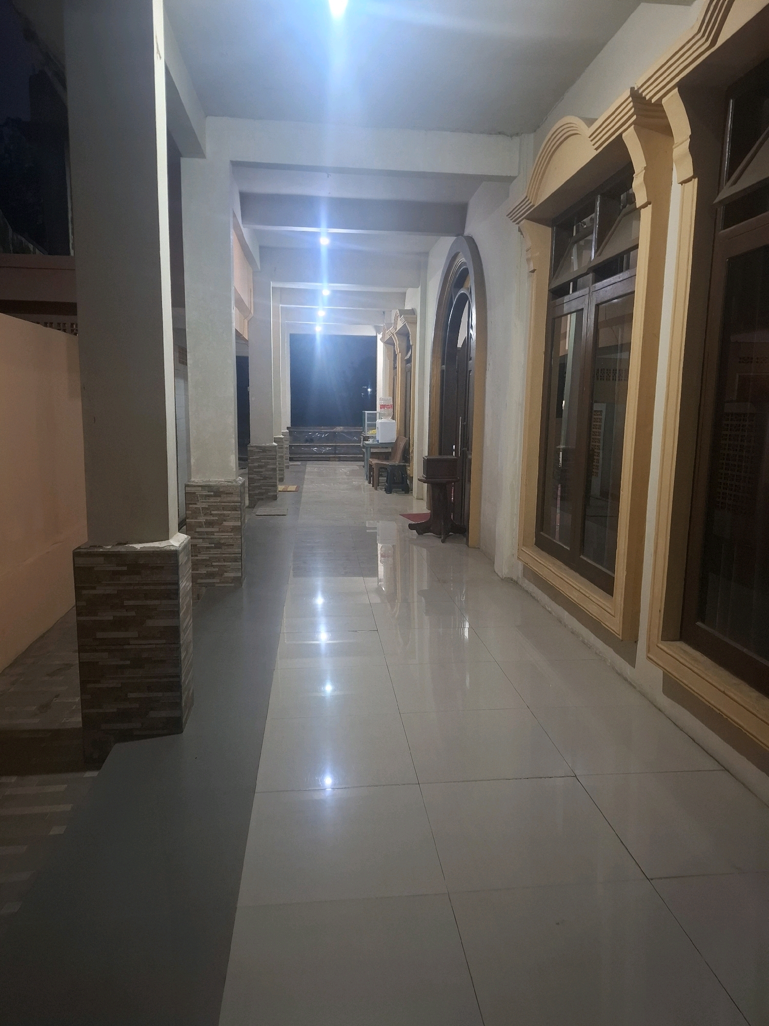 This image depicts an outdoor hallway or walkway at night, possibly part of a building or complex in Ciheulang Tonggoh, Indonesia. The walkway is illuminated by artificial lighting, creating reflections on the glossy tiled floor. On the left side, there are large pillars with stone tile accents. The right side of the walkway features large windows with ornate wooden frames. In the background, the hallway extends into darkness, with a few dimly lit areas suggesting further rooms or an outdoor space. There are no people visible in the image. The overall scene conveys a sense of quiet and possibly an establishment that is closed for the night.