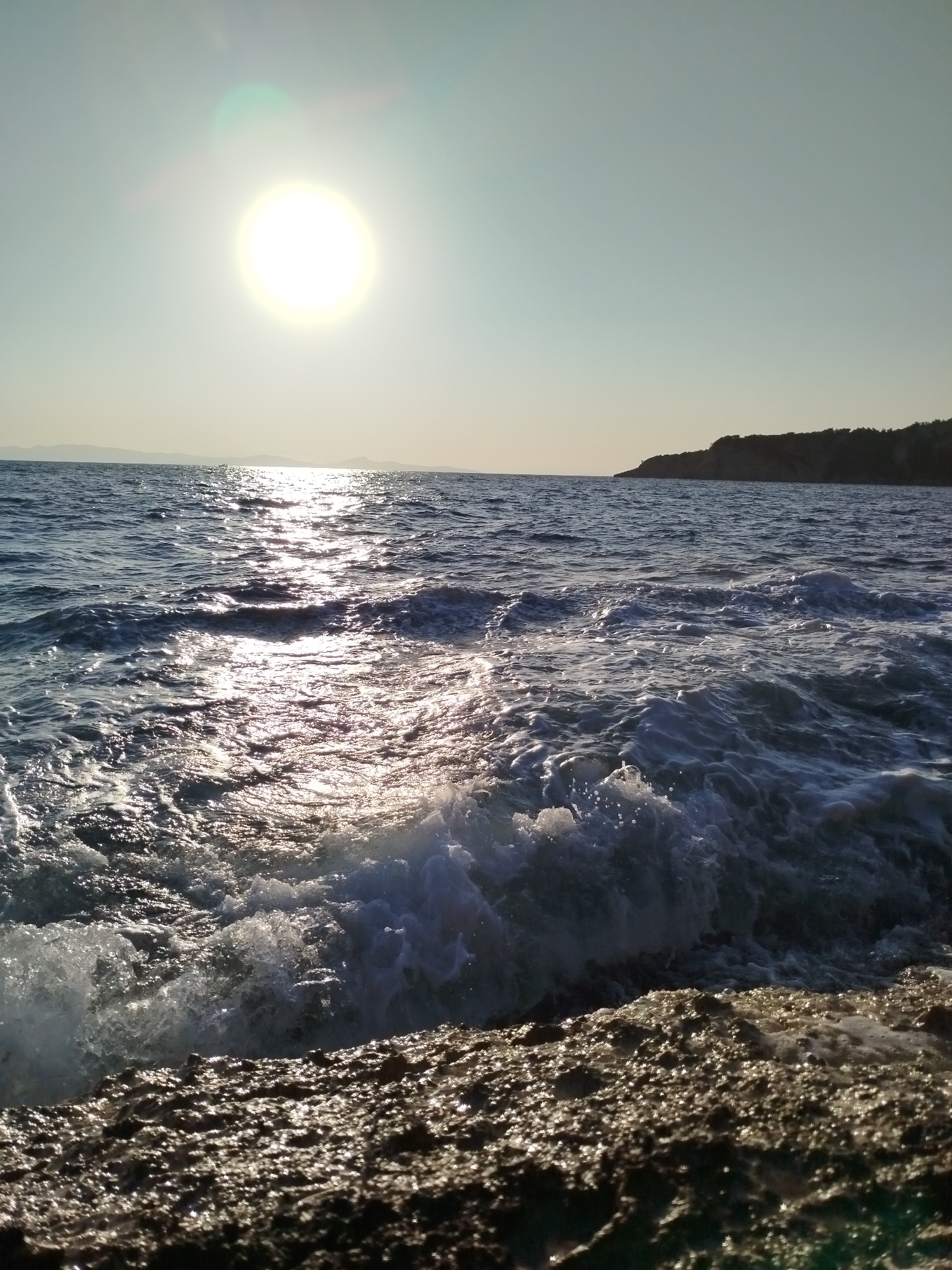 A bright sun shines in the upper left quadrant of the image, casting a glare on the sea. The sun's rays reflect off the rippling water, creating a shimmering effect. Waves crash against the rocky shore in the foreground. In the distance, a dark, tree-covered hill slopes down to the sea. The sky is clear and pale blue. The image appears to be taken from the coast of Karaburun, Turkey, a peninsula known for its rugged coastline and clear waters.