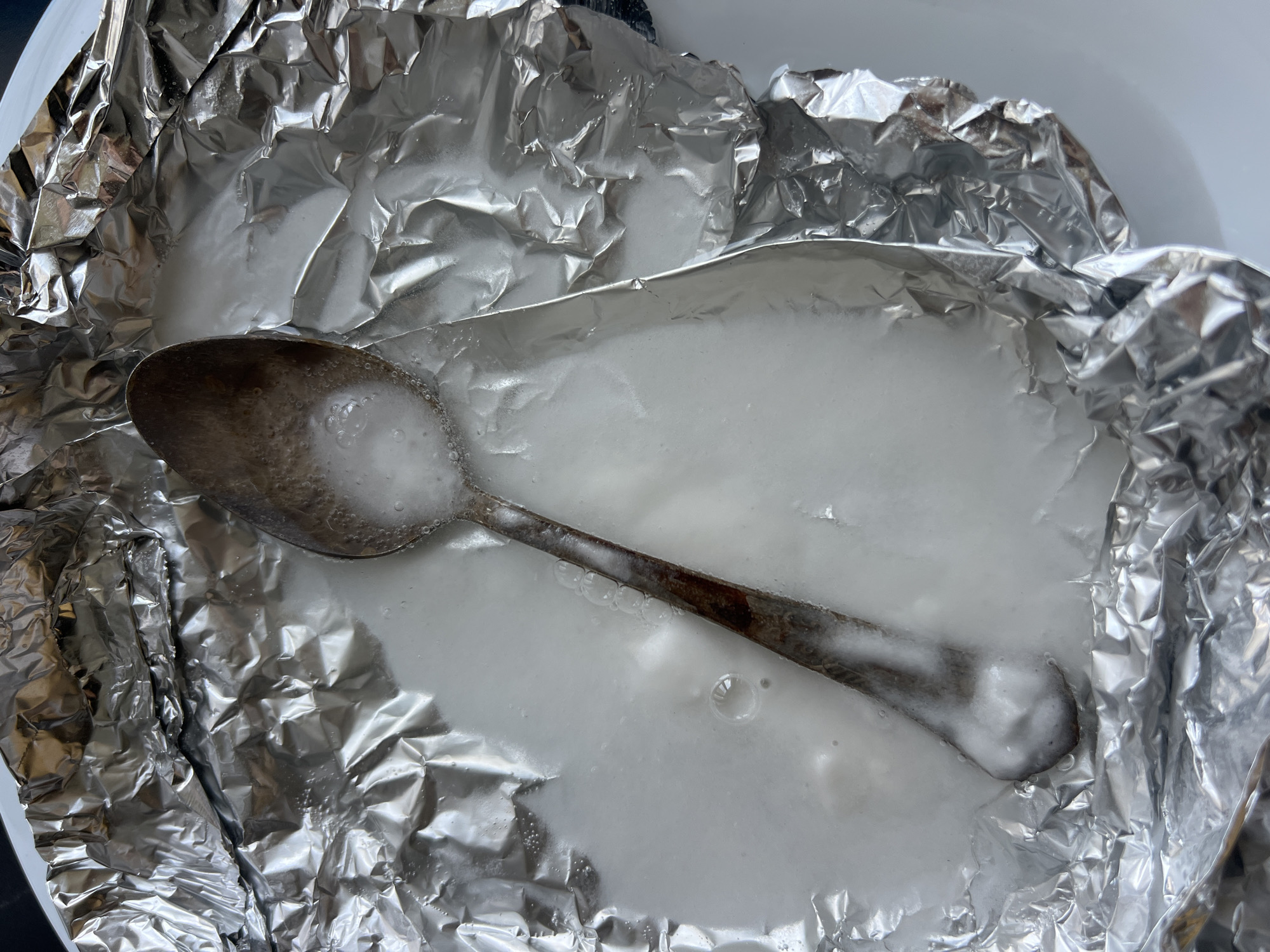 The image shows a close-up, top-down view of a metal spoon resting in a white, frothy liquid contained within crumpled aluminum foil. The foil is shaped into a makeshift dish and appears to be on a white plate. Bubbles are visible in the liquid and on the surface of the spoon, indicating a chemical reaction or effervescence. The spoon itself shows signs of wear and possibly tarnish. The lighting is bright, causing reflections on the foil. The scene is indoors. There is no discernible context indicating a location in Sochi, Russia, or any specific activity beyond the spoon being in a frothy substance within aluminum foil.