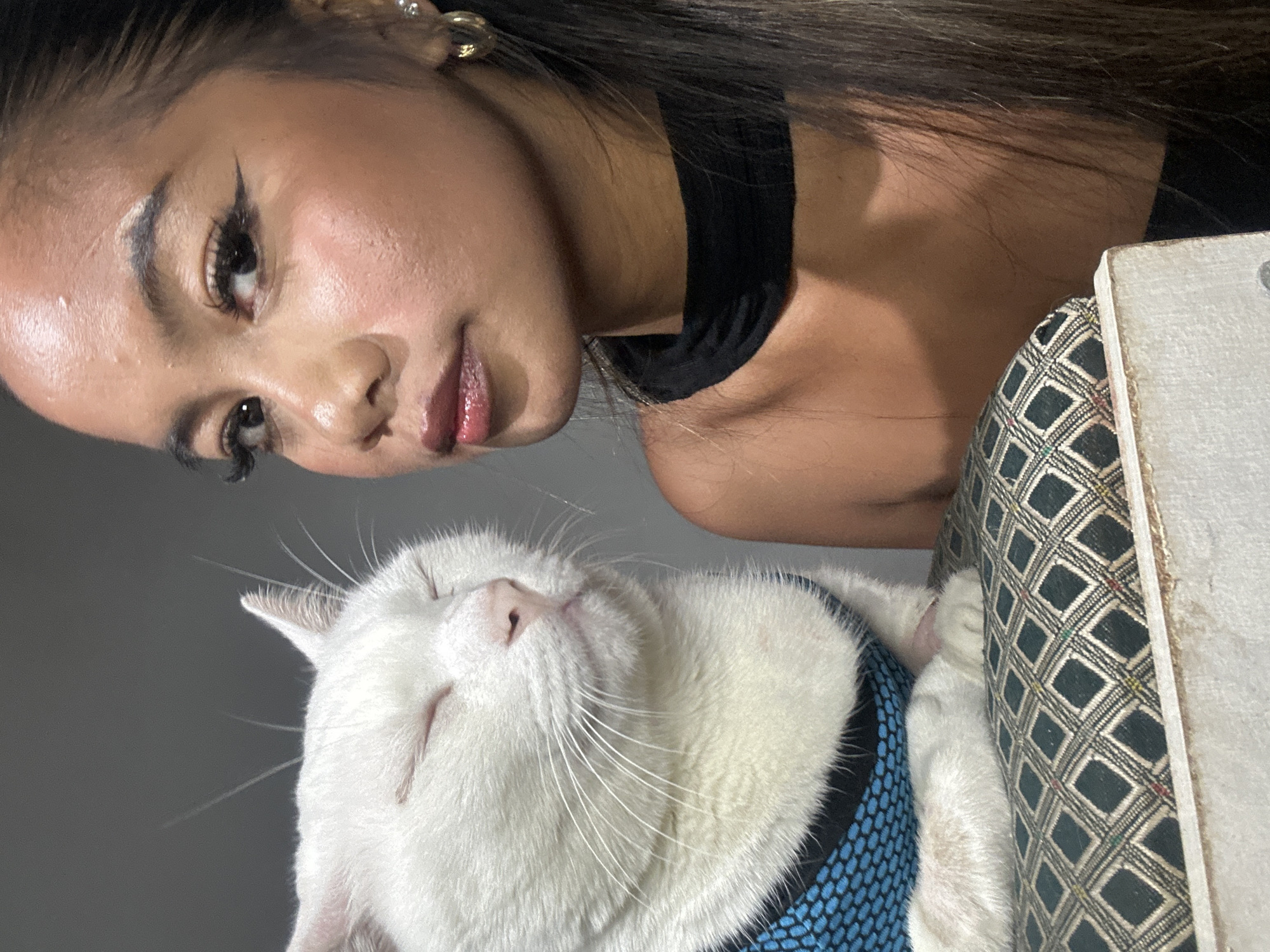 A woman with long, dark hair wearing a black choker and gold earrings is shown above a white cat with closed eyes. The woman is looking at the camera with a slight smile. The cat is wearing a blue harness. The cat and woman are resting against a cushion with a checkered pattern and a white border. The location is likely indoors. There is no indication of the specific location within North Jakarta, Indonesia.