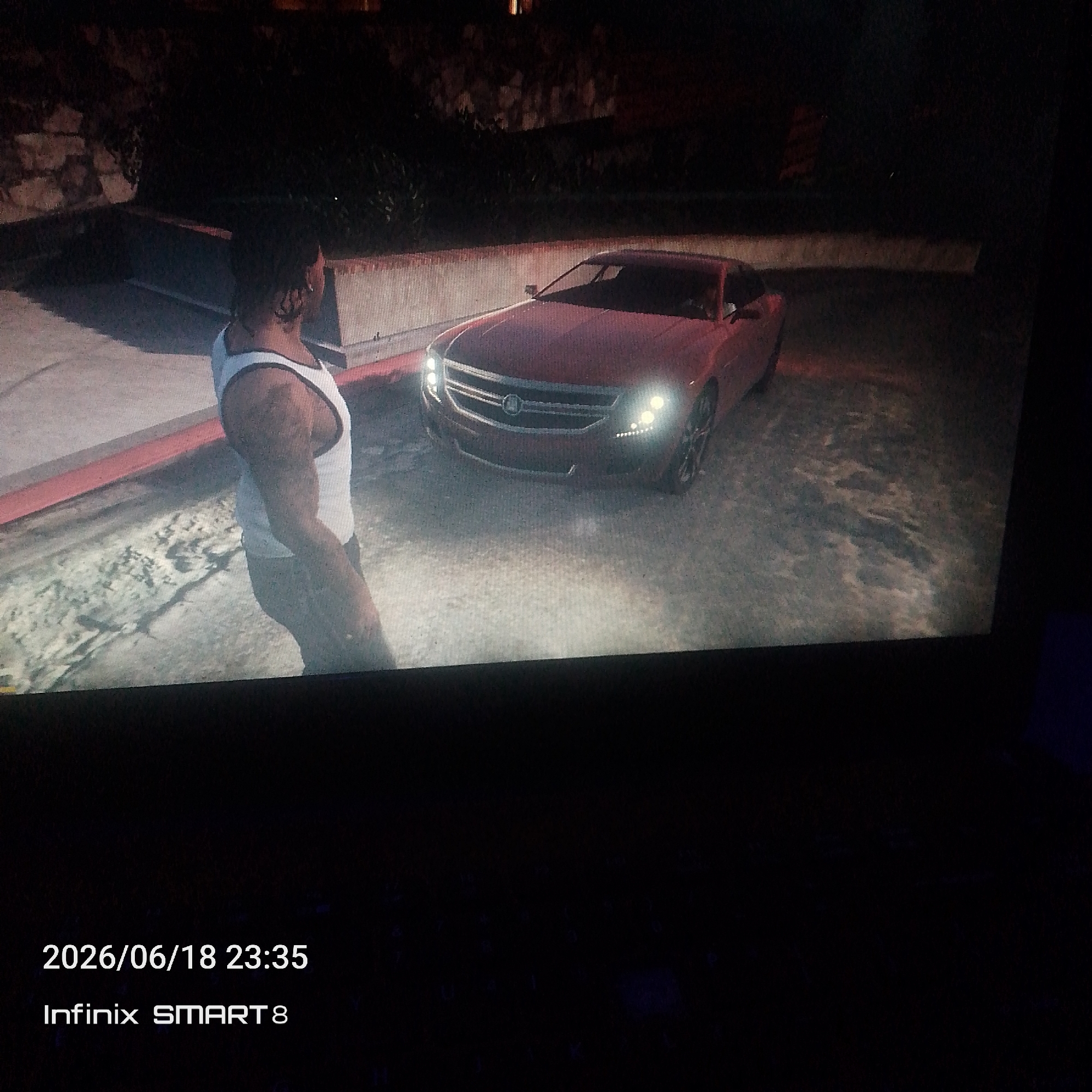 A man wearing a white tank top stands next to a red car. The car has its headlights on, illuminating the scene. The environment appears to be an urban setting at night, with a stone wall and foliage visible in the background. The man is facing away from the camera, and his arms are at his sides. The car is parked on a paved surface, possibly a driveway or street. The image also includes a timestamp and device information at the bottom: "2026/06/18 23:35 Infinix SMART 8".
