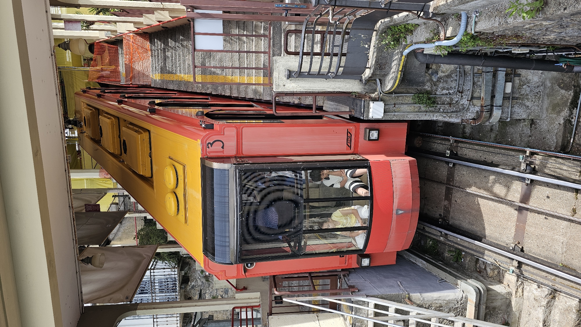 The image depicts the funicular railway car in Brunate, Italy. The railway car is red and yellow, with the number "3" prominently displayed on its side. Passengers are visible inside the car, including two people in the front window. The railway car is on tracks that run along a concrete pathway. Adjacent to the tracks is a concrete wall with exposed pipes and vegetation. A staircase is next to the railway car at the station. A protective barrier is present. The environment appears to be urban and mountainous.