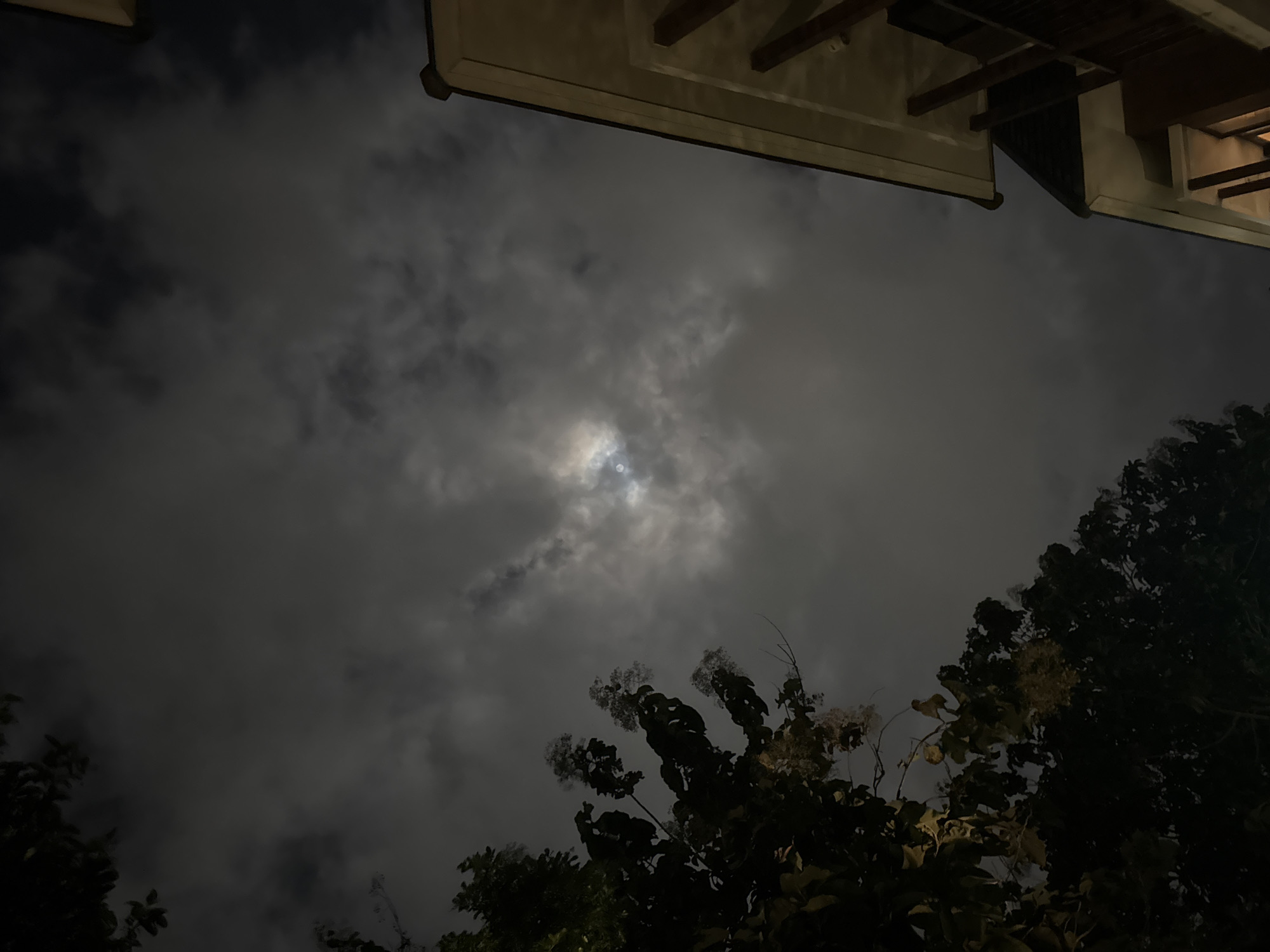 The media file presents an upward view of a cloudy night sky, featuring a partially obscured moon as the central light source. The moon's glow emanates from behind dense clouds, creating a luminous, diffuse patch of light in the middle of the frame that illuminates the surrounding cloud formations.
In the lower right and bottom-left portions of the image, dark silhouettes of tree branches and foliage are visible. The trees appear to be robust, with some lighter clusters, possibly flowers or lighter leaves, noticeable on the branches in the lower right.
The upper section of the image shows the underside of an architectural structure or roof overhang. This includes visible wooden beams, eaves, and a soffit or ceiling, all rendered in shadows but subtly highlighted by the ambient moonlight.
The scene takes place at night, evident from the dark sky and the moon's prominence. The weather is cloudy, with the moon peeking through, creating an atmospheric and somewhat mysterious ambiance. There is no human activity or visible text present. The composition captures a tranquil natural scene juxtaposed with subtle man-made elements, suggesting a quiet, nocturnal setting.