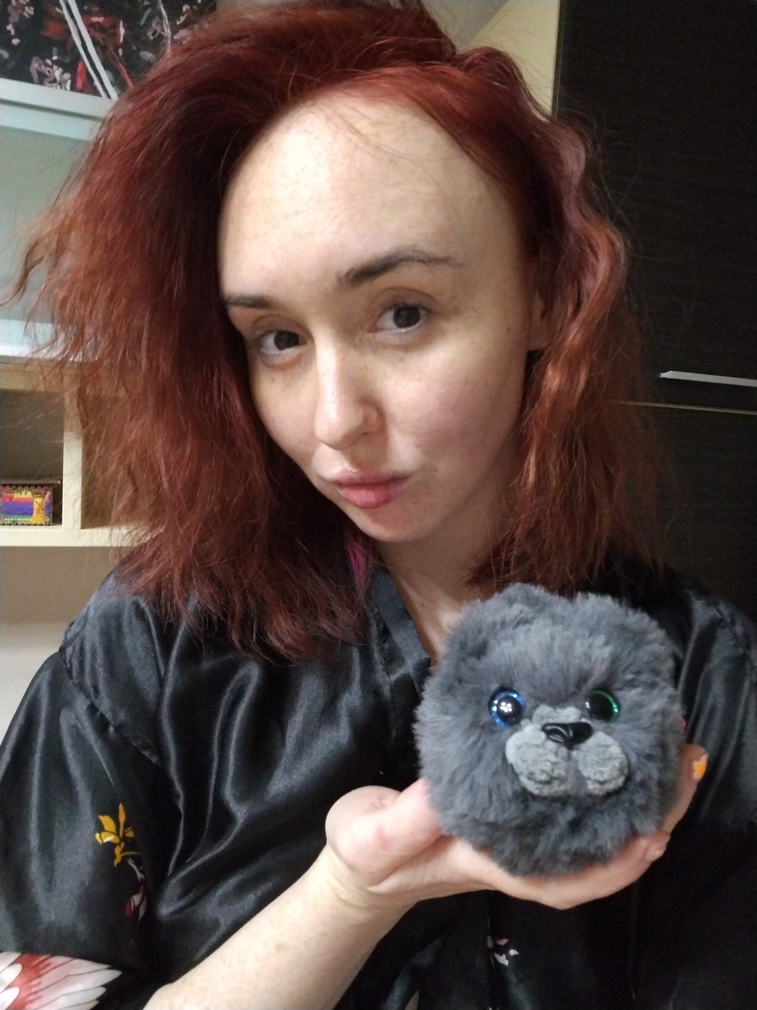 The image displays a woman indoors in Izhevsk, Russia. She has fair skin, reddish-auburn hair, and is looking directly into the camera with a slight pout. She is wearing a dark, possibly black, satin robe with a yellow and red pattern visible on the left sleeve. Her right hand, with orange nail polish on a visible finger, holds a small, spherical, grey plush toy. The toy features one blue eye and one green eye, a black nose, and a light grey stitched muzzle area. The background includes a light-colored shelving unit on the left with a small, multicolored object, and a dark brown or black furniture surface on the right. A portion of a poster or artwork is visible above the shelving unit.