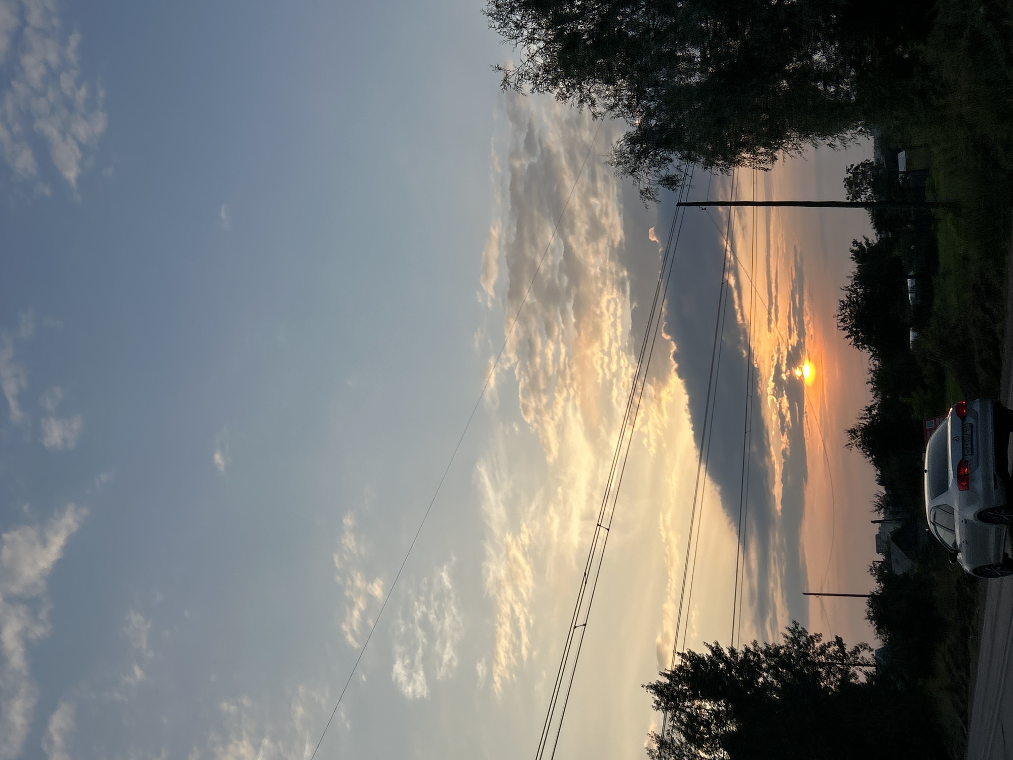 The image depicts a street scene in Chaikovsky, Russia, during a sunset. A silver sedan is driving on a road in the foreground, with its rear facing the camera. Power lines are strung across the sky, with a utility pole visible. Trees and some houses are silhouetted against the sky, which is illuminated by the setting sun. The sun is partially obscured by clouds, casting a warm orange and yellow glow across the horizon. The sky is a pale blue with scattered white clouds. The overall scene suggests a tranquil, late afternoon or early evening atmosphere.