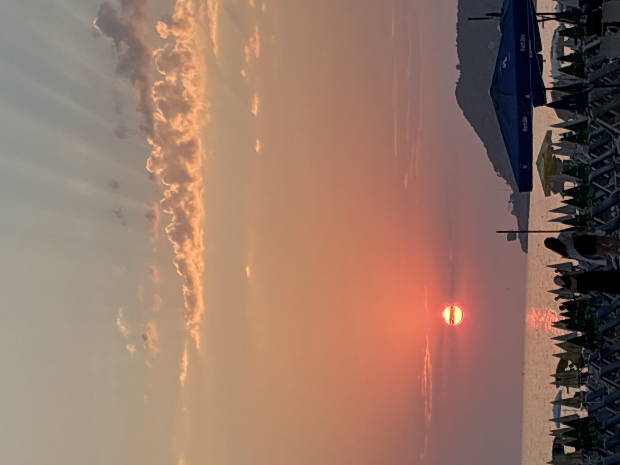 This image depicts a sunset scene at Cleopatra Beach in Alanya, Turkey. The sun, a vibrant orange orb, is partially obscured by a thin layer of clouds and is low on the horizon, casting a warm glow across the sky and sea. Rays of sunlight are visible streaming upwards from behind the clouds. The water of the Mediterranean Sea appears calm, with a reflection of the sun visible on its surface.
In the foreground, rows of beach chairs and umbrellas are arranged, some of which are closed. A few people are visible, walking on the beach. One person is wearing a dark dress and a headscarf, while another person, further to the right, is wearing a white top. The beach is lined with numerous green and blue umbrellas. In the background, a dark, mountainous silhouette of a hill or island rises from the sea. Several blue beach umbrellas with the text "#artibir" are prominent in the mid-ground. The overall atmosphere is serene and characteristic of an evening at a beach resort.