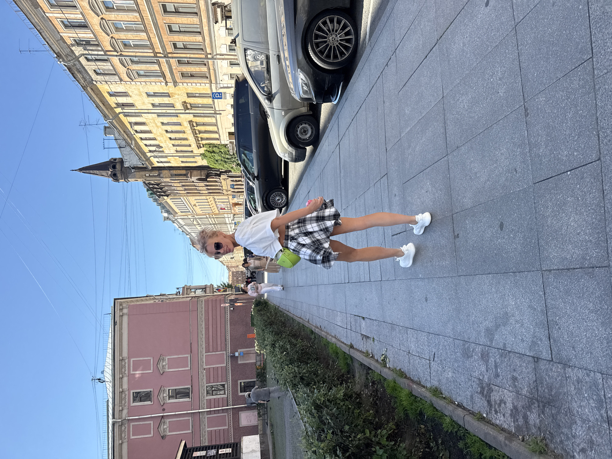 In Saint Petersburg, Russia, a young woman with blonde hair styled in an updo, wearing sunglasses, a white crop top, a black and white plaid skirt, and white sneakers, walks away from the camera down a paved sidewalk. She carries a bright green purse on her left shoulder. The sidewalk is lined with a green hedge on the left and parked cars on the right. The buildings in the background are a mix of historical architecture, including a pink building with ornate white trim and a tall, dark spire on one of the buildings. The sky is clear and blue. The overall scene depicts a sunny day in a European city.