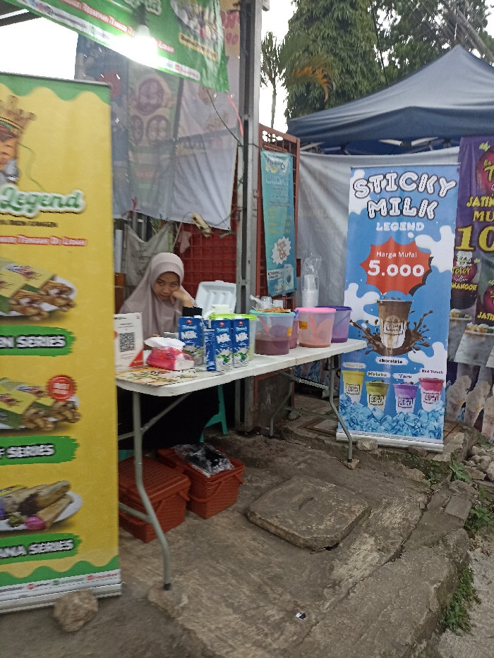 A young woman in a headscarf sits at a table in front of a stall in Jatinangor, Indonesia, located at coordinates -6.935, 107.773. She is wearing a white headscarf with black trim and a long-sleeved, white shirt. She has her chin on her hand. In front of her is a table with various items, including: four cartons of milk, two plastic containers of what appears to be red liquid, two plastic containers of what appears to be blue liquid, a white paper napkin dispenser, a white paper bag, a red plastic container, and a white plastic container. The table is a folding table with a white top and silver legs. The stall appears to be selling drinks called "Sticky Milk Legend", and the price is advertised at 5,000. There is a large blue and white sign behind the table advertising the drink. The background of the image includes an awning, a wall, and a fence. There are two plastic storage bins behind the table. The ground is paved and cracked. The sky is overcast.