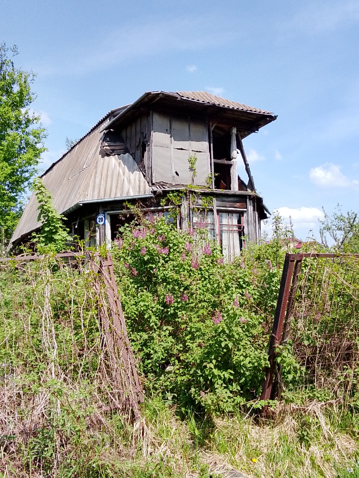 The image shows an abandoned, dilapidated house in Снопок Новый, Russia. The structure has a dilapidated roof, damaged walls, and overgrown vegetation obscuring the front. A rusty metal fence surrounds the property, which is overgrown with weeds and bushes. A blue and white number "20" is visible on the house. The sky is blue with scattered clouds. There are no people visible in the image.