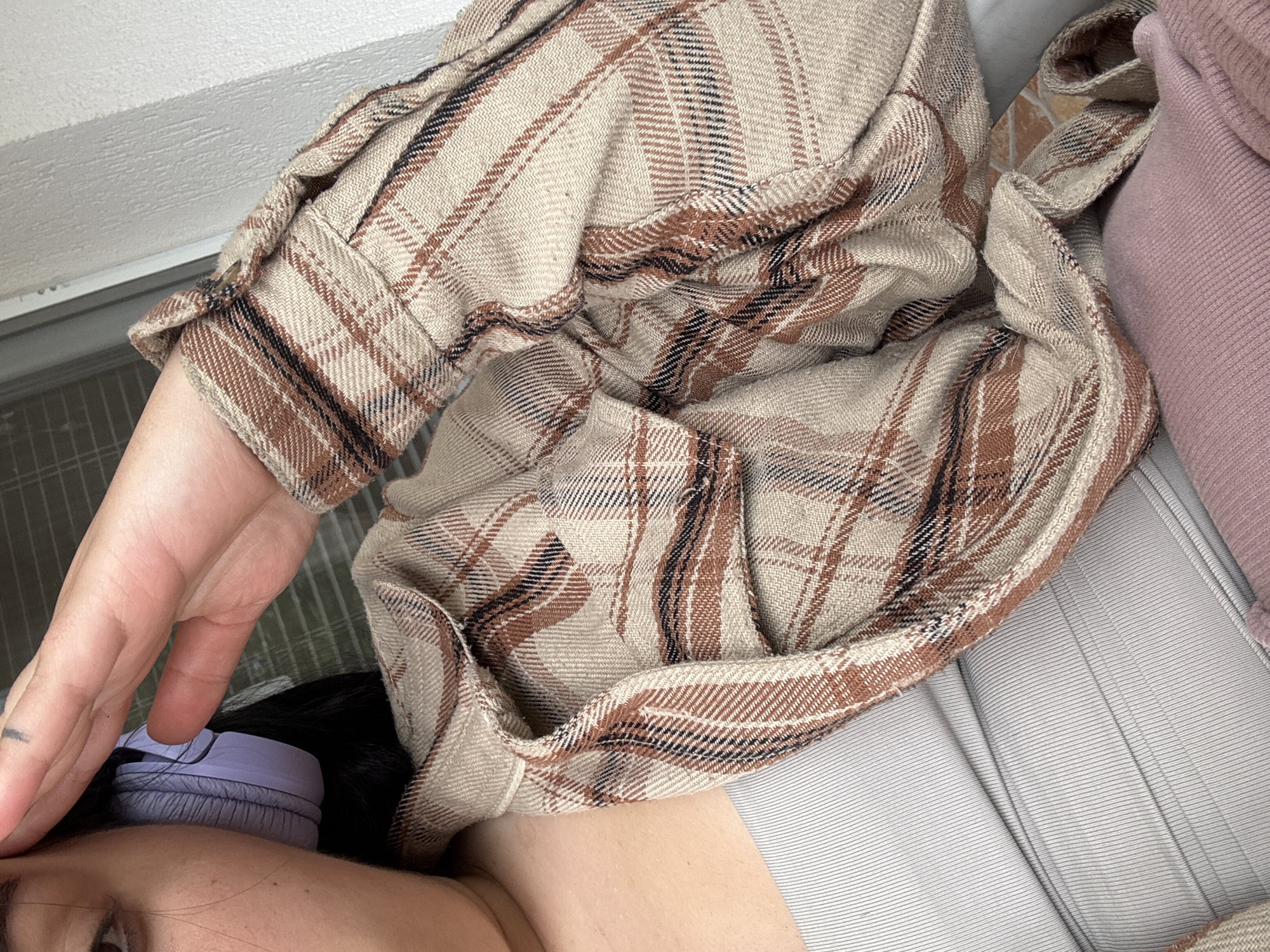 The image depicts a person wearing a plaid flannel shirt, lying down. A hand is visible near the person's face, which is partially obscured. Purple headphones are positioned near the person's ear. The person is lying on a light grey, vertically striped surface with pink fabric near the top right corner. The photo seems to be taken indoors. There is no information present to indicate the location is in Asenovgrad, Bulgaria.