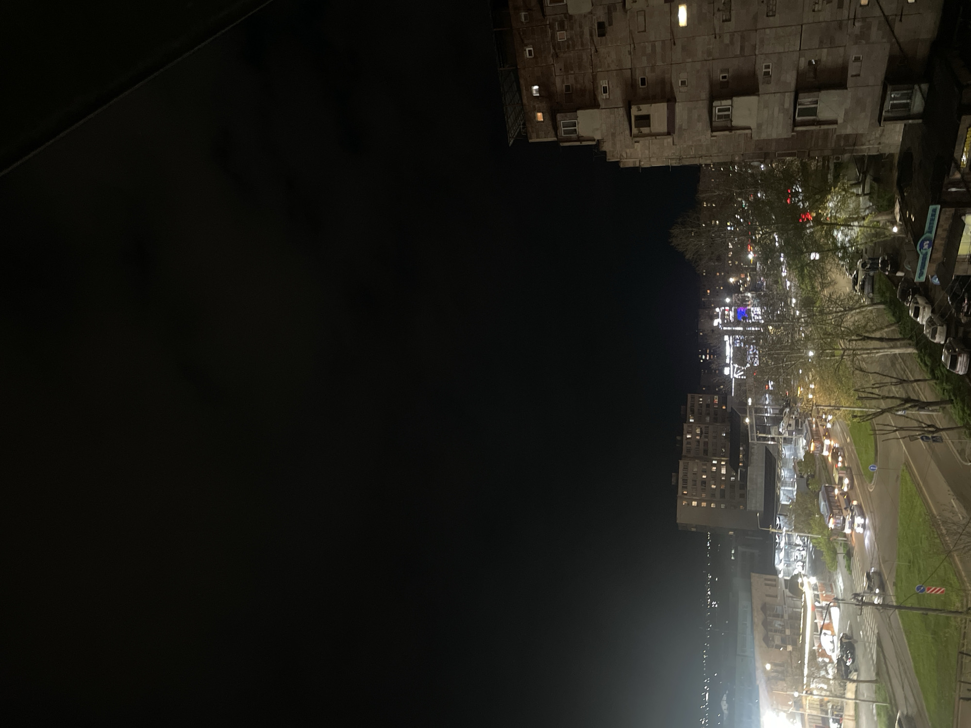 This nighttime photograph, taken from an elevated perspective, captures a bustling urban street scene in Yerevan, Armenia. The overall impression is one of a city alive with artificial light against a dark sky.
In the upper part of the frame, a dark, angular surface occupies the top left, suggesting the edge of a building or a window frame from the vantage point of the photographer. To the right, a prominent, multi-story apartment building with a facade of numerous windows dominates the midground. Many windows emit warm, yellow light, indicating occupied units. The building's architecture appears functional and blocky.
Below this building, a wide city street stretches across the lower and central parts of the image. It's busy with traffic, visible as trails of white headlights and red taillights from numerous cars. The street is bordered by bare trees, possibly indicating late autumn or early spring, and strips of grass. Streetlights cast a bright glow on the road surface and surrounding areas. In the distance, a panorama of city lights extends, suggesting further urban development.
Notable details include a blue sign with white text on the right side of the street, likely belonging to a commercial establishment, though the text is not decipherable. A red and white striped sign, possibly a road warning, is also visible. The time of day is clearly night, with the only illumination coming from artificial sources. No individual people are distinctly visible, but the active traffic suggests ongoing city life.
