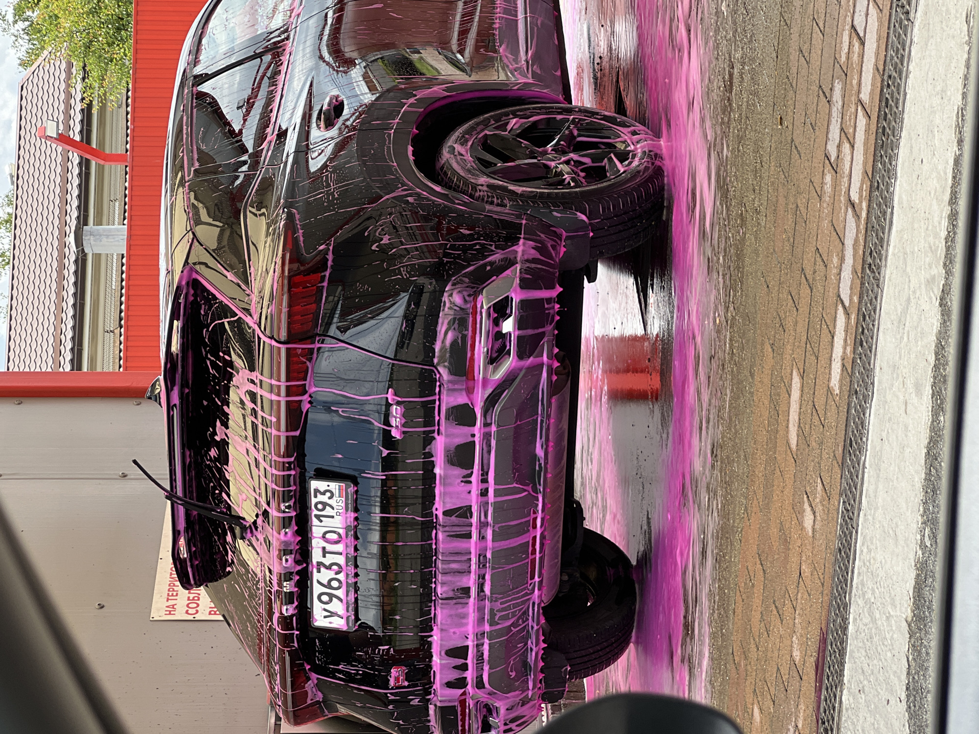 A black SUV is being washed at an outdoor car wash station. The vehicle is covered in bright pink foam, which is dripping down the sides and pooling on the ground. The car has a Russian license plate that reads "У963ТО 193". In the background, there is a red fence and a building with a tiled roof. A sign in Russian is visible on the wall behind the car. The ground is paved with reddish-brown bricks. The scene appears to be a typical car washing service in Krasnodar, Russia. There are no people visible in the image.