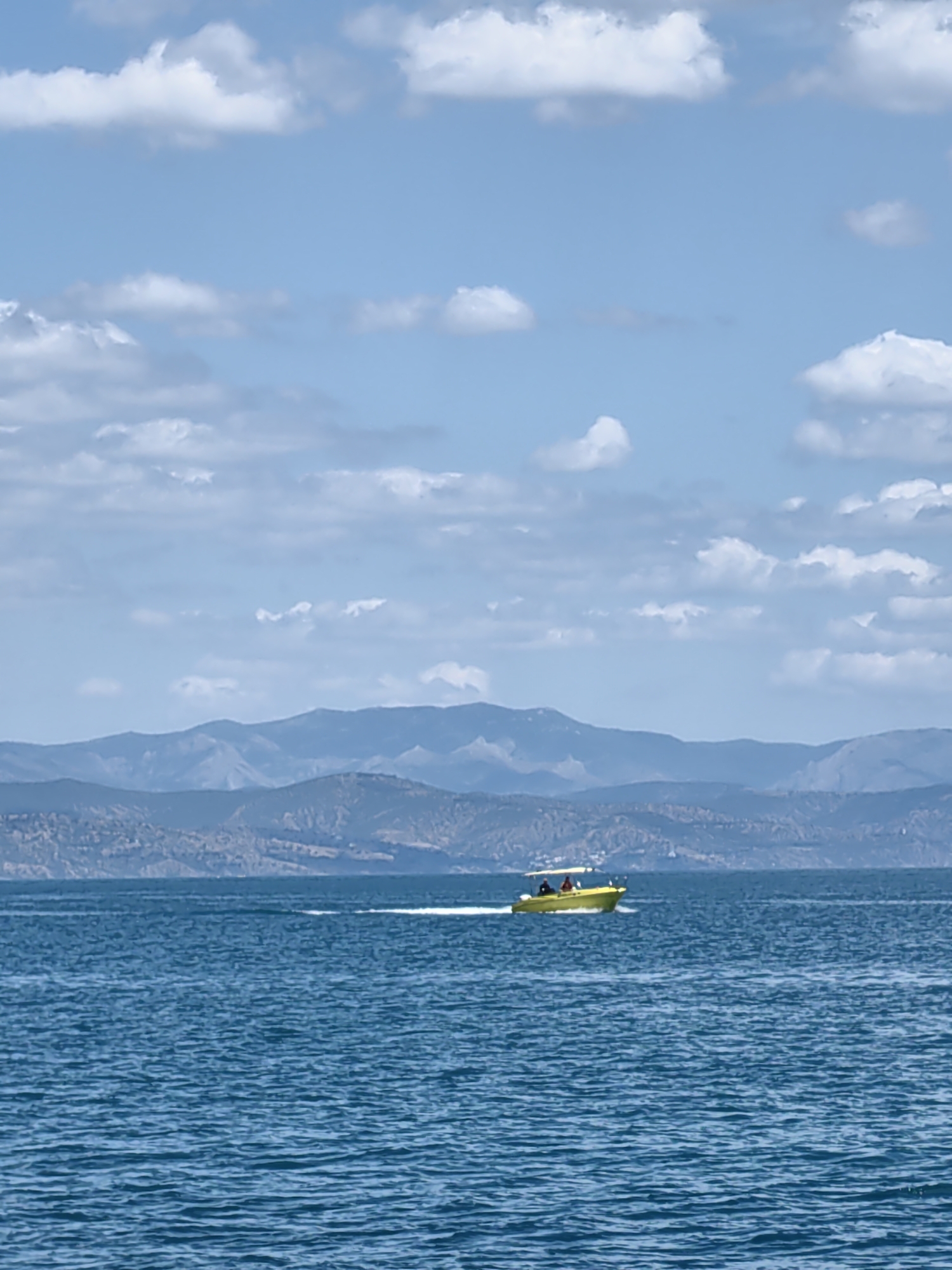 A yellow motorboat with two occupants is navigating across the choppy, blue waters of the sea. The boat is positioned in the middle ground, leaving a white wake in its path. In the background, a range of hazy, blue-toned mountains stretches across the horizon, suggesting a coastal landscape. The sky above is a pale blue, dotted with scattered white clouds. The overall scene is tranquil and suggests an outdoor recreational activity.
