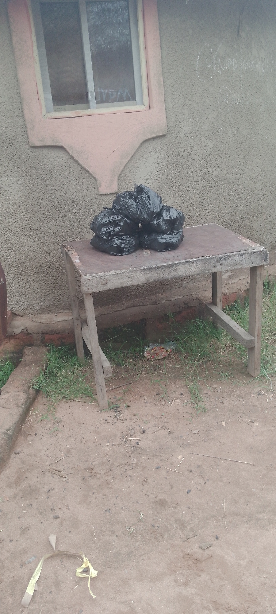 The image depicts an outdoor scene in Jalingo, Nigeria, featuring a roughly constructed wooden table with a dark, possibly metal, top. On the table rests a pile of black garbage bags. Behind the table, a textured, light-colored wall is visible, with a window frame that appears to be painted pink and yellow. The ground in front of the table is a dirt surface with sparse grass. A piece of yellow ribbon or tape lies on the ground in the lower left corner of the image. Graffiti is visible on the wall to the right of the window.