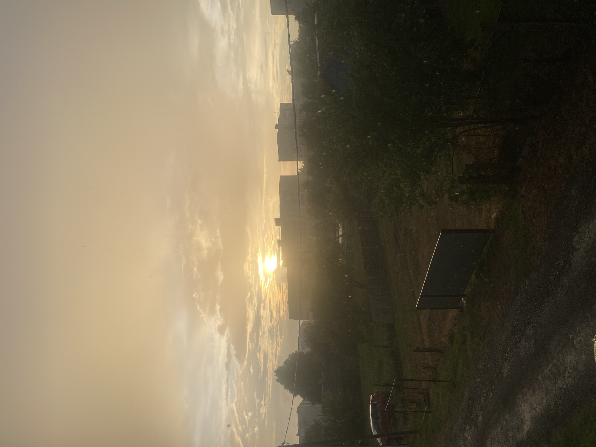 This is a photograph of a rural or semi-urban landscape in Krupki, Belarus, captured during a rain shower at sunset or sunrise. The sky is dominated by warm, golden hues, with the sun visible as a bright, diffused orb through the clouds and rain. Scattered white clouds are present, and the atmosphere appears hazy, likely due to the rain and low light conditions.
In the foreground, a gravel path is visible, wet from the rain, with some puddles reflecting the sky. To the left of the path, a grassy area is present, bordered by a simple fence. A red car is parked to the left of this area. Further into the scene, a line of trees and bushes forms a green barrier. Beyond the trees, several multi-story apartment buildings are visible, characteristic of Soviet-era architecture, suggesting a residential area. A wooden fence runs across the mid-ground, separating different plots of land. Small white specks, indicative of falling raindrops, are scattered throughout the image, particularly visible against the darker trees and sky. Power lines are also present, stretching across the frame. The overall impression is one of a somewhat overcast, moody, and tranquil scene, with nature and urban development coexisting.
