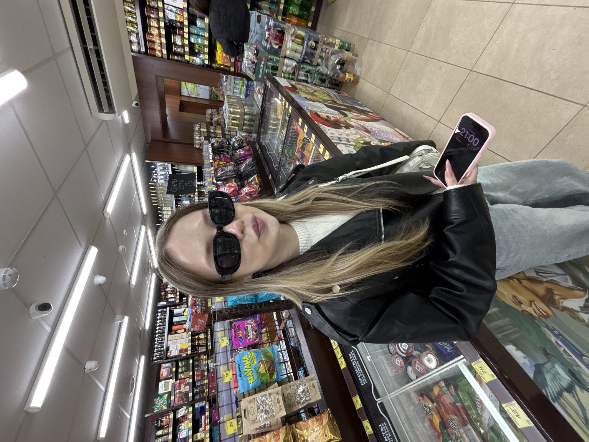 A young woman is captured in a vertical, waist-up shot inside what appears to be a convenience store or small supermarket in Фоки, Russia. She has long blonde hair, is wearing black sunglasses, a black leather jacket, and a white sweater, with light-colored pants visible. She holds a pink-cased smartphone in her right hand, displaying the time "21:00," indicating it is 9 PM. Her expression, partially obscured by sunglasses, seems neutral.
The background reveals typical store elements: brightly lit shelves stocked with various packaged goods, including snacks like "MAYAMA" and "Pencil" candies, and beverages like "Apostel Brau" beer. Refrigerated display cases line the aisle, with one showing "Mil" (likely milk products) and another with prices like "79" and "42" (presumably rubles). Overhead, bright fluorescent lights illuminate a tiled ceiling, where an air conditioning unit and several small surveillance cameras are mounted.
Visible text on store signage and product labels includes "ПРОДУКТЫ МОЛОЧНОГО ЖИРА" (Dairy Products) and a partially visible word "Оремя" (likely "Время" meaning Time) on a sign, possibly indicating opening hours or just displaying the time. Prices such as "26", "42", "59", and "79" are also noticeable on shelf tags. The overall scene suggests a casual late-evening visit to a local store.